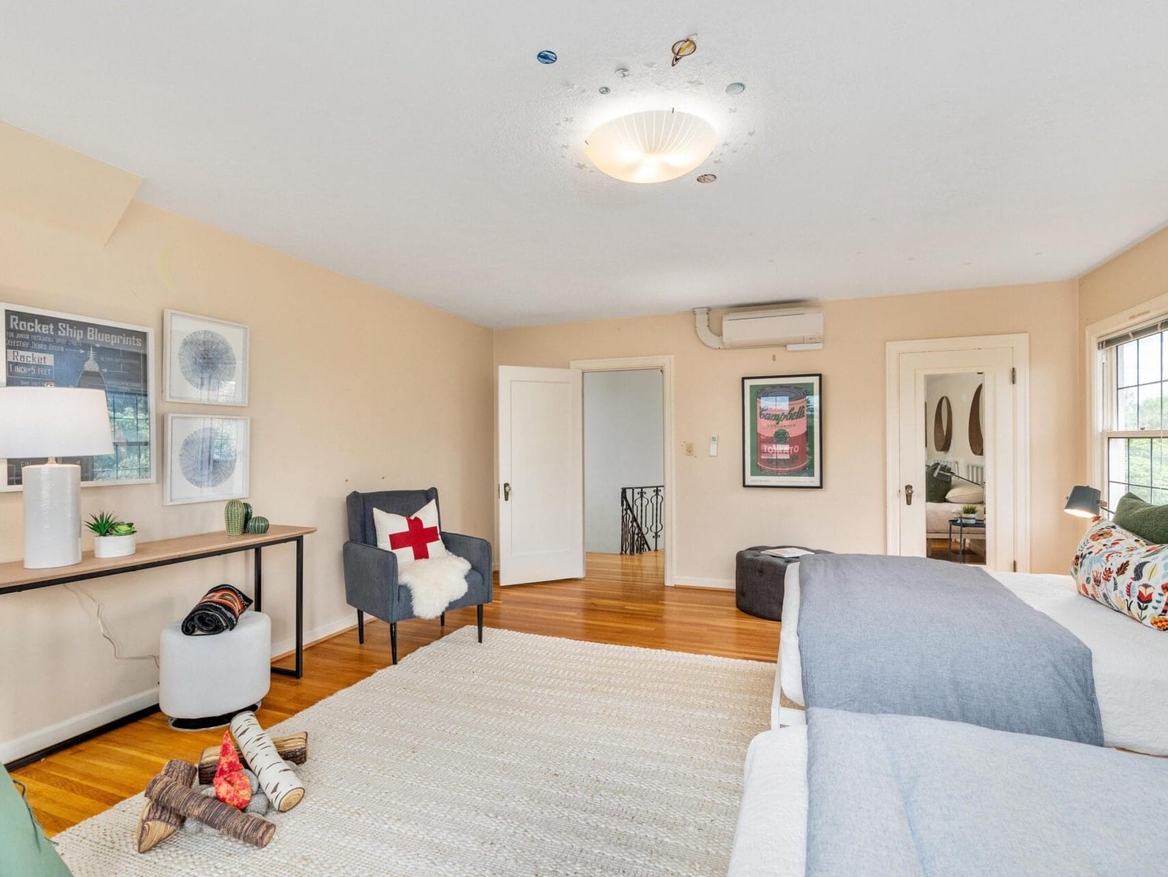 A cozy bedroom with two beds, a soft beige carpet, and a wooden floor. The room features a cushioned chair with a red cross pillow, a table with a lamp, and decorative artworks on the walls. Windows let in natural light.