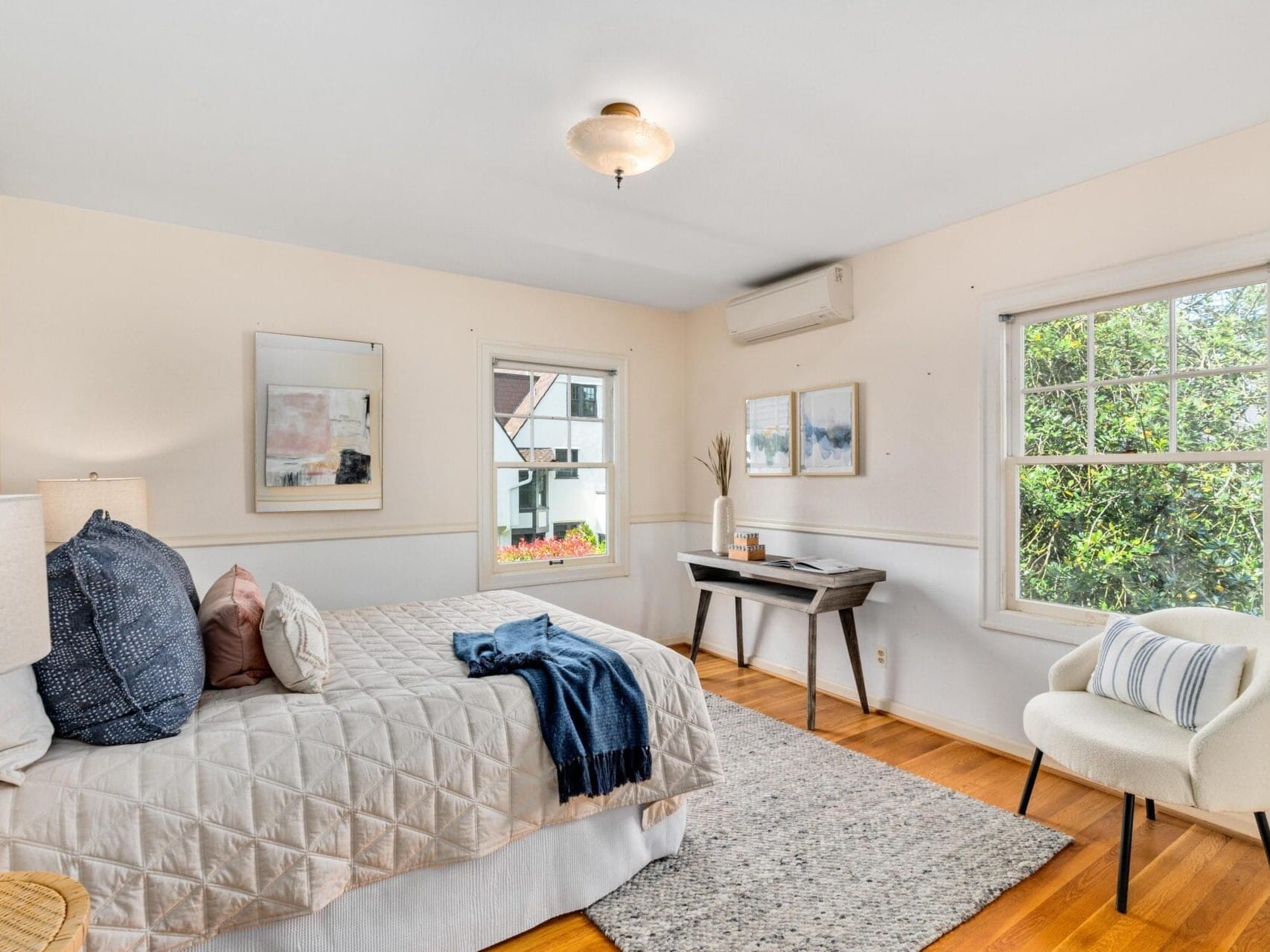 A cozy bedroom with a neatly made bed featuring a white quilt and blue cushions. Theres a light-colored armchair and a small wooden table by the window. The room is bright with natural light, and a green view is visible outside.