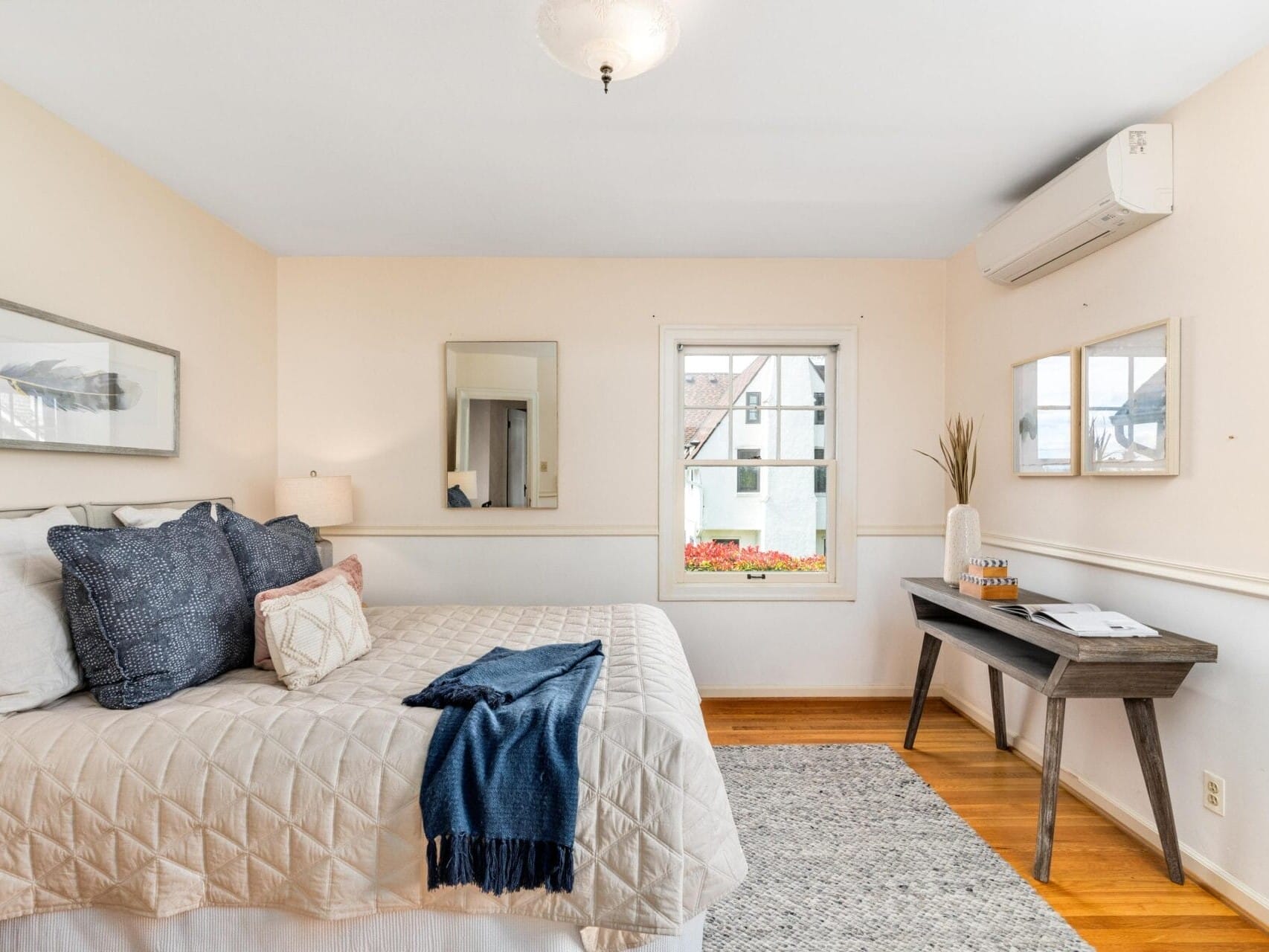 A cozy bedroom featuring a neatly made bed with blue and beige pillows and a blue throw. The room includes a wooden desk with decor, an air conditioning unit, and a window offering a view of the outdoors. Mirrors and framed art adorn the light-colored walls.