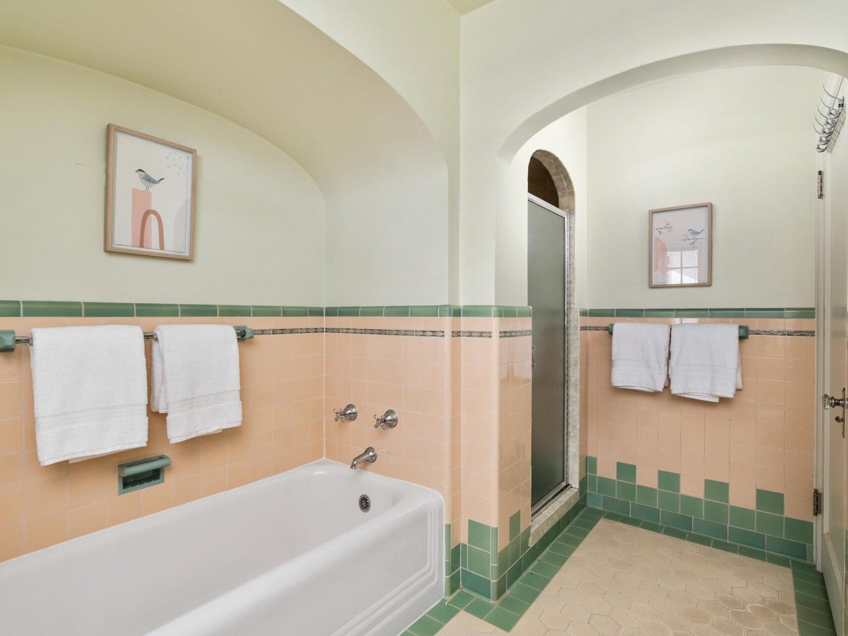 A vintage-style bathroom with pastel green and peach tiles, featuring a white bathtub, silver faucets, and two hand towels on a rack. Artwork with bird motifs decorates the walls, and a separate shower area is visible in the background.