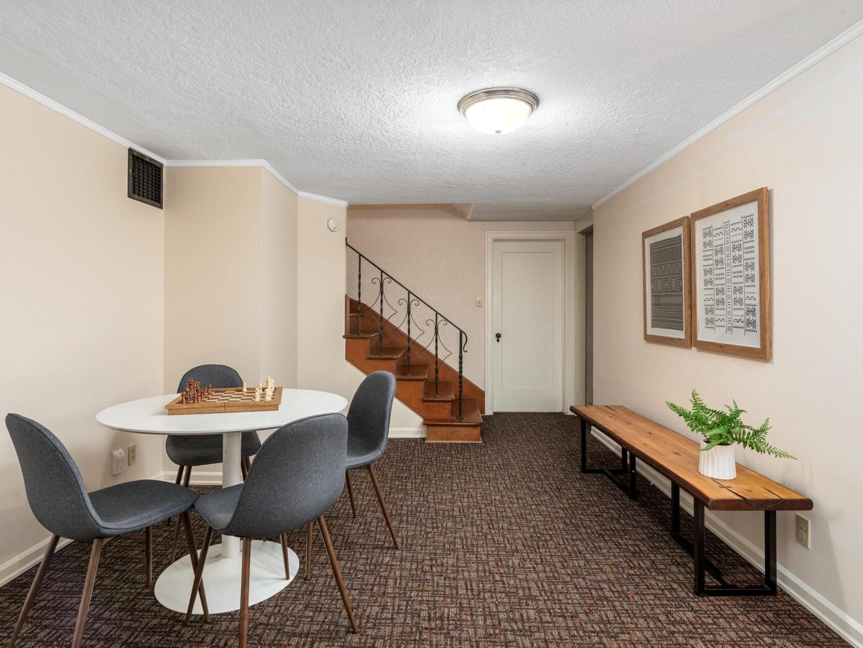 A small room features a round white table with a chess set and four gray chairs. A wooden bench with a potted plant is against one wall. A staircase with a wrought iron railing leads to a closed door. Two framed artworks adorn the beige walls.