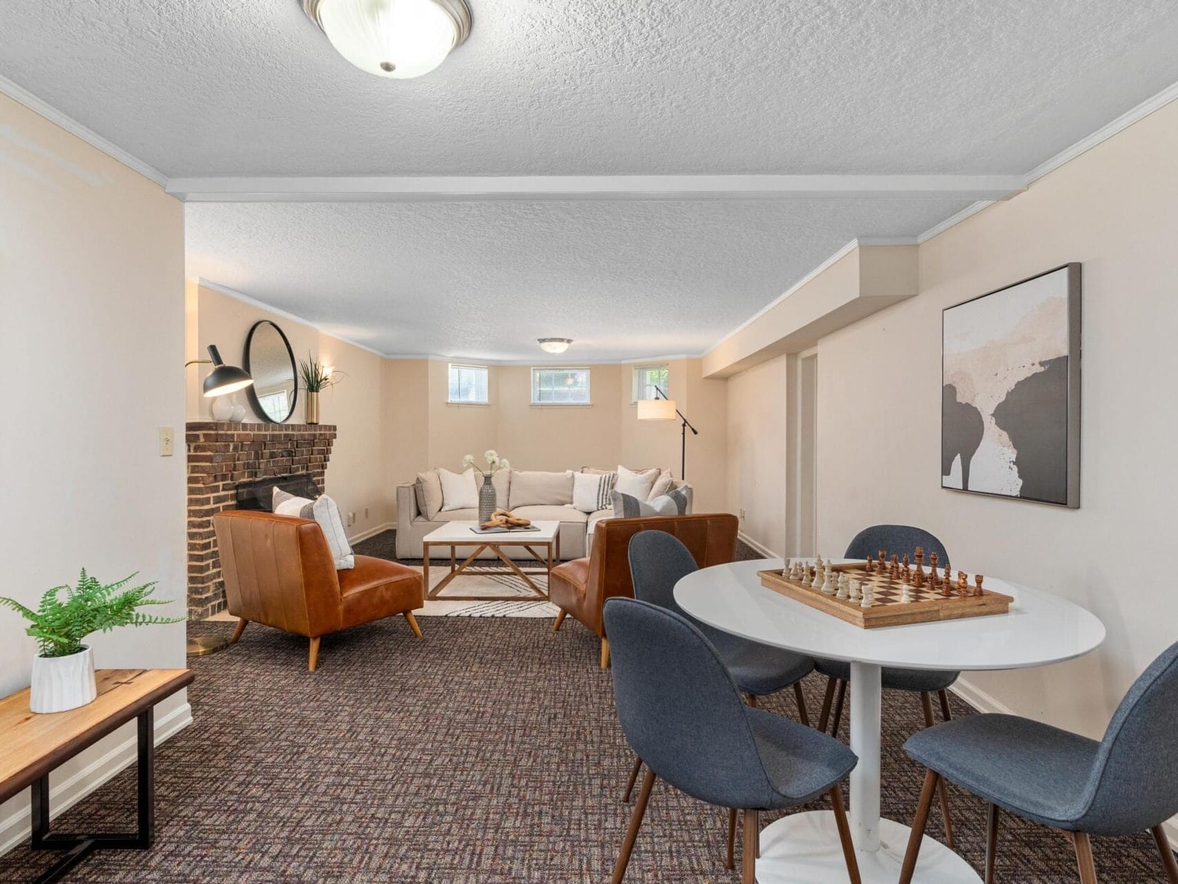 A cozy living room with a brick fireplace, two brown leather chairs, and a beige sectional sofa. A round table with a chessboard is in the foreground, surrounded by four dark chairs. Soft lighting and abstract art decorate the walls.