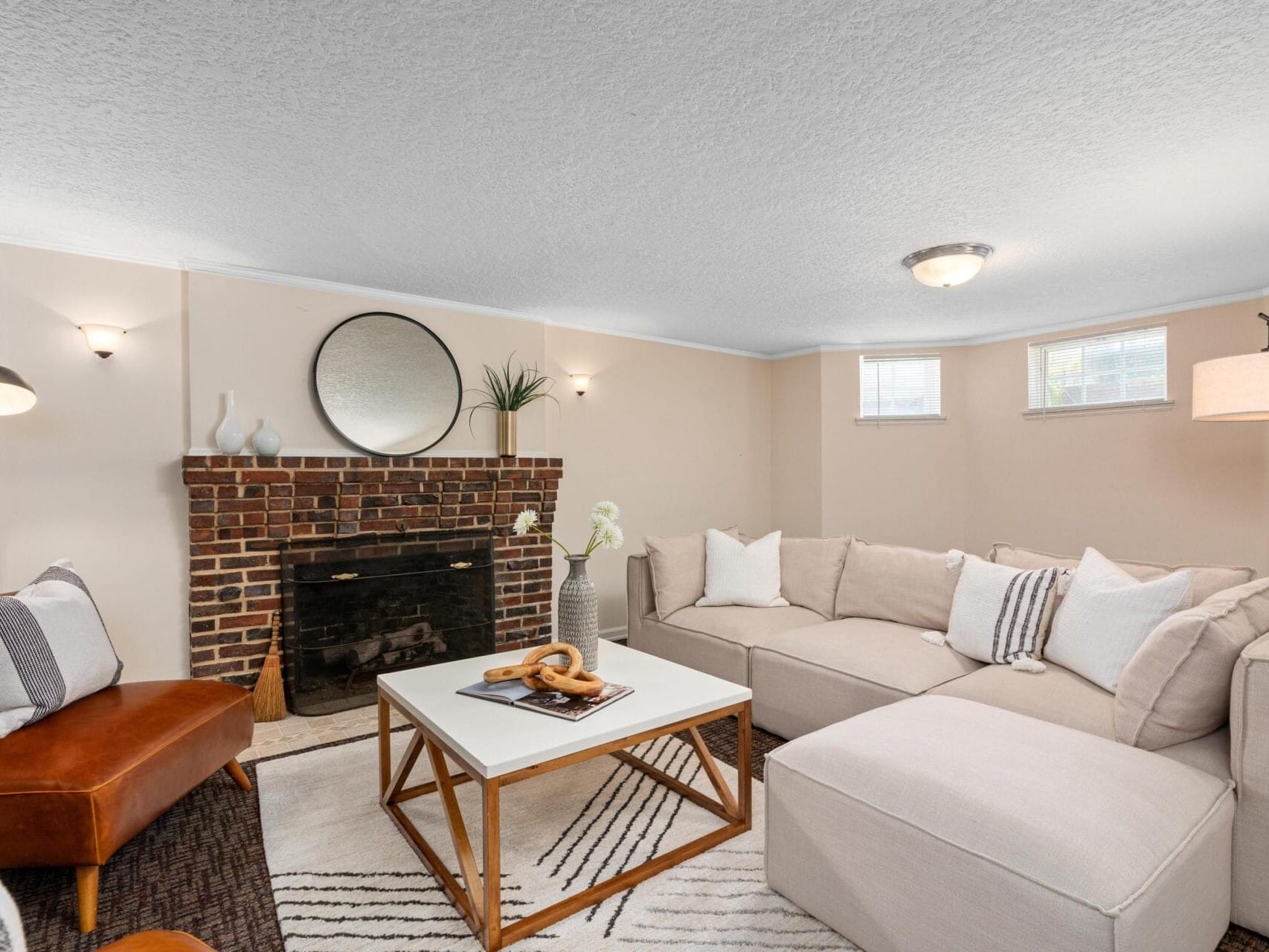 A cozy living room with a brown leather chair, a beige sectional sofa, and a white coffee table on a patterned rug. A brick fireplace with a round mirror above it is adorned with vases and a plant. Soft light filters through small windows.