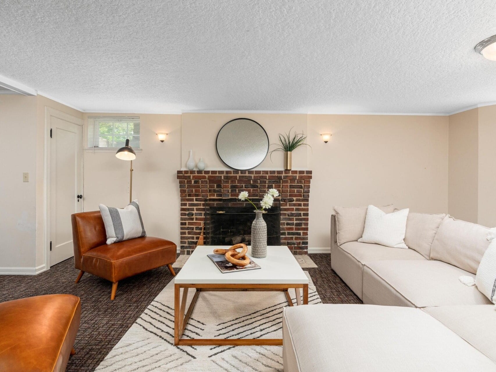 A cozy living room with a beige sectional sofa and two brown leather chairs. A white coffee table with decor sits on a patterned rug. A brick fireplace is adorned with a round mirror and plant. Soft lighting illuminates the neutral walls.