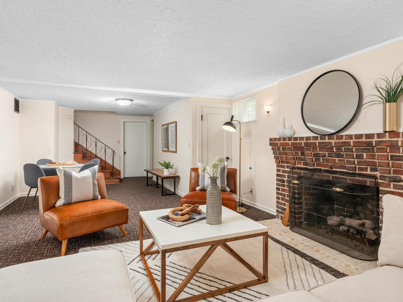 A cozy living room featuring a brick fireplace with a round mirror above it. The room includes a white rug, a beige couch, two brown leather chairs, a white coffee table with decor, and a dining area visible in the background.