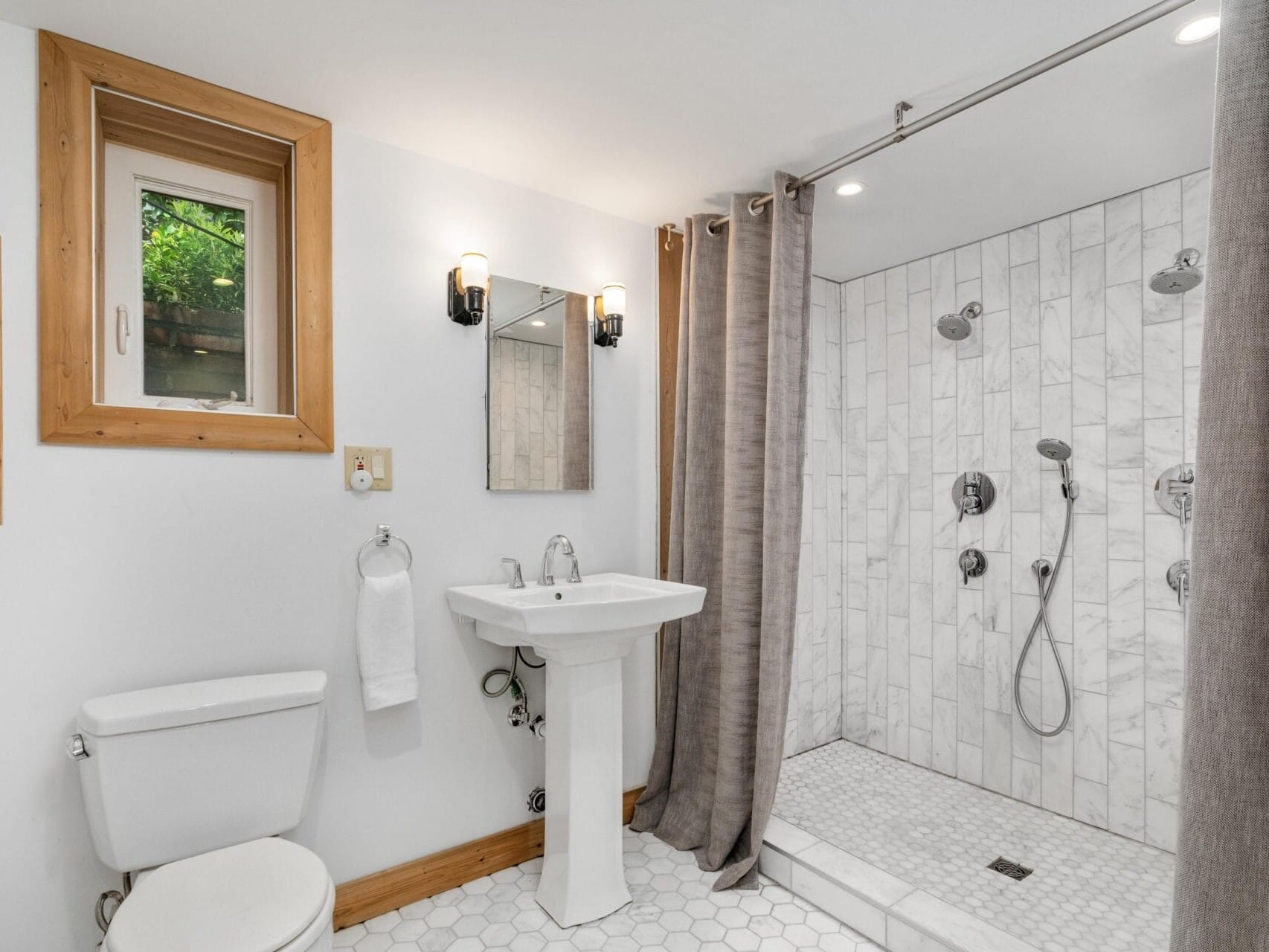 Modern bathroom with a walk-in shower featuring white tiles and dual shower heads. A pedestal sink with a square mirror and two wall sconces is adjacent to a white toilet. A small window and hexagonal floor tiles complete the clean design.