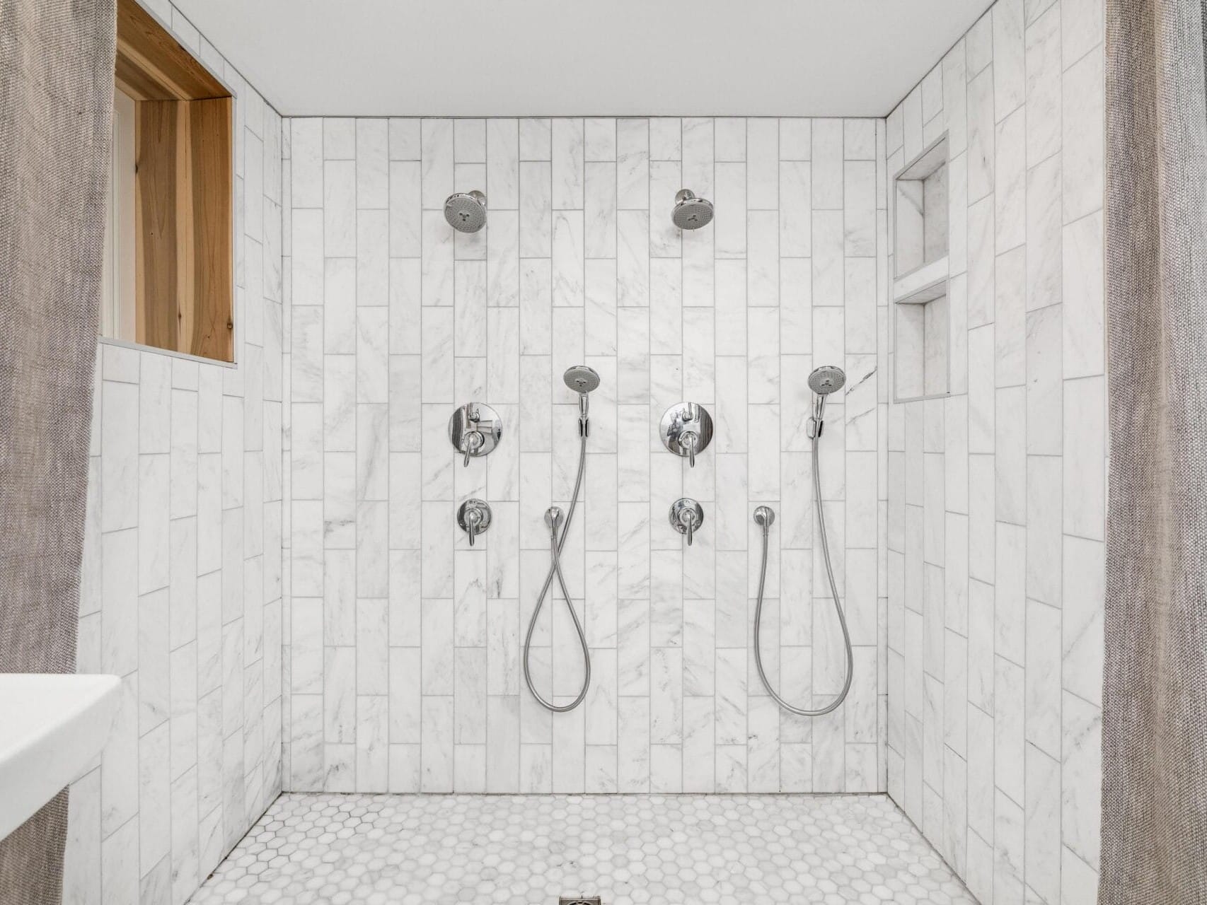 A modern shower with two showerheads and handheld attachments. The walls are lined with white marble tiles, and the floor features small hexagonal tiles. There is an open cubby for storage and a wooden-framed window on the left wall.