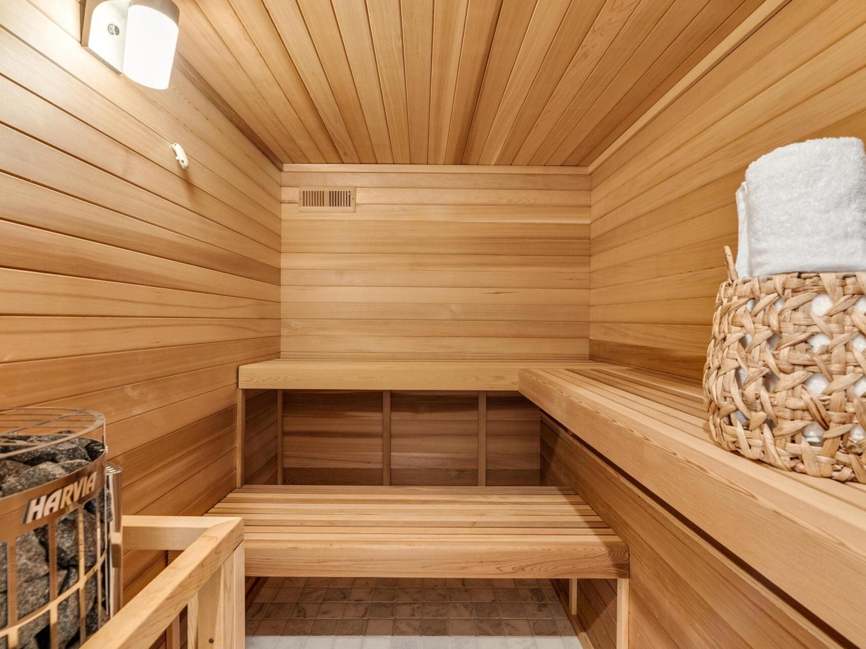 A wooden sauna room with a bench along the walls. A basket filled with white towels sits on the bench. A sauna heater with sauna rocks is in the corner. The walls and ceiling are lined with smooth light wood planks.