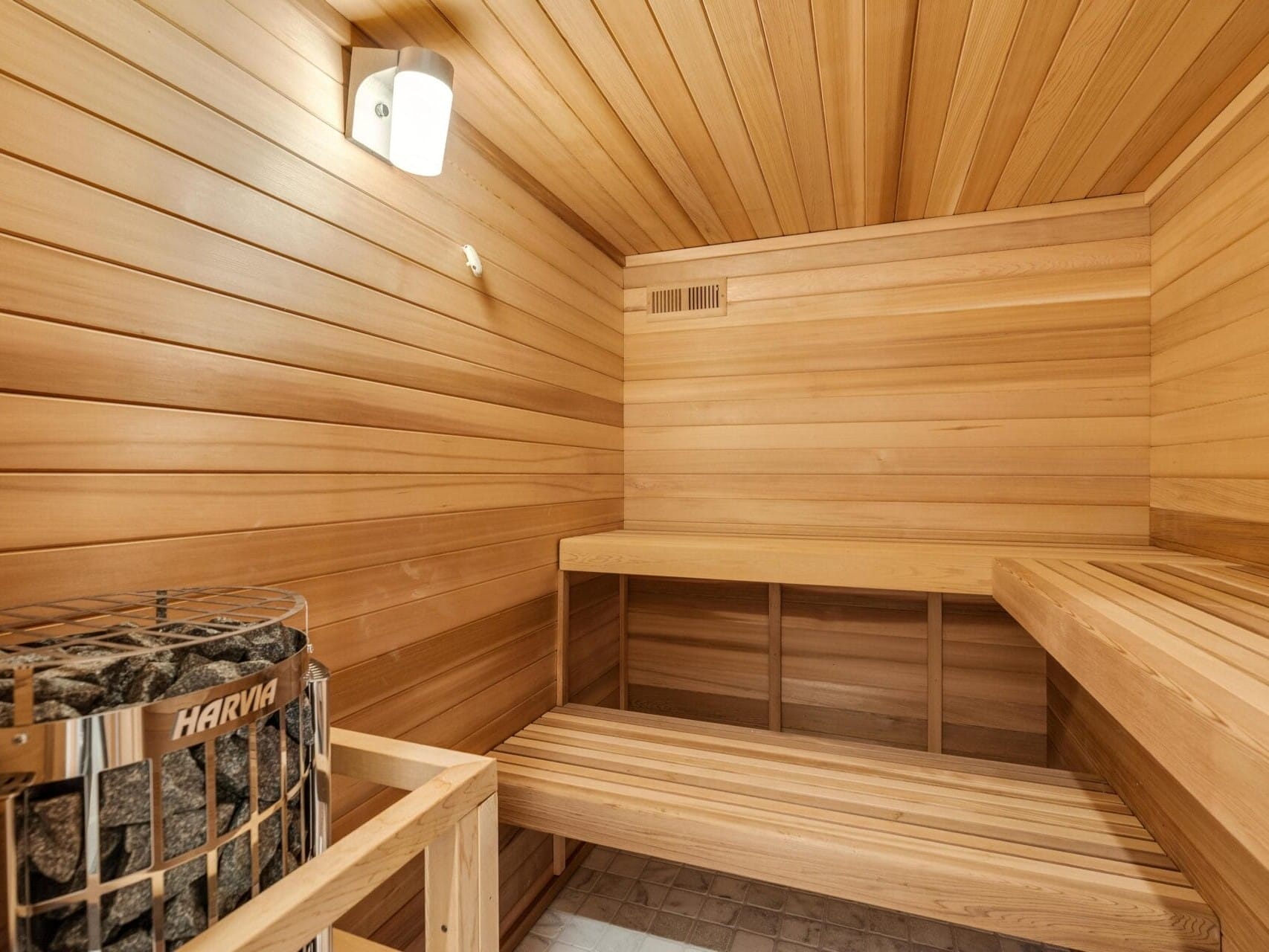 A wooden sauna interior featuring a bench on the right, a wall-mounted light fixture, and a Harvia stove in the corner filled with sauna stones. The walls and ceiling are designed with smooth wooden panels.