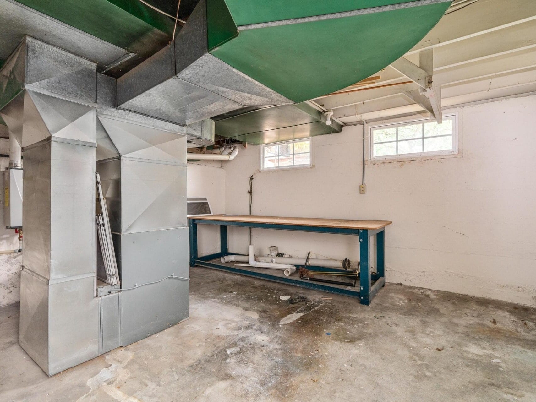 A basement room with exposed ceiling beams, ductwork, and a furnace. There’s a large workbench along the wall beneath two small windows. The floor is concrete, and pipes are visible near the bench.