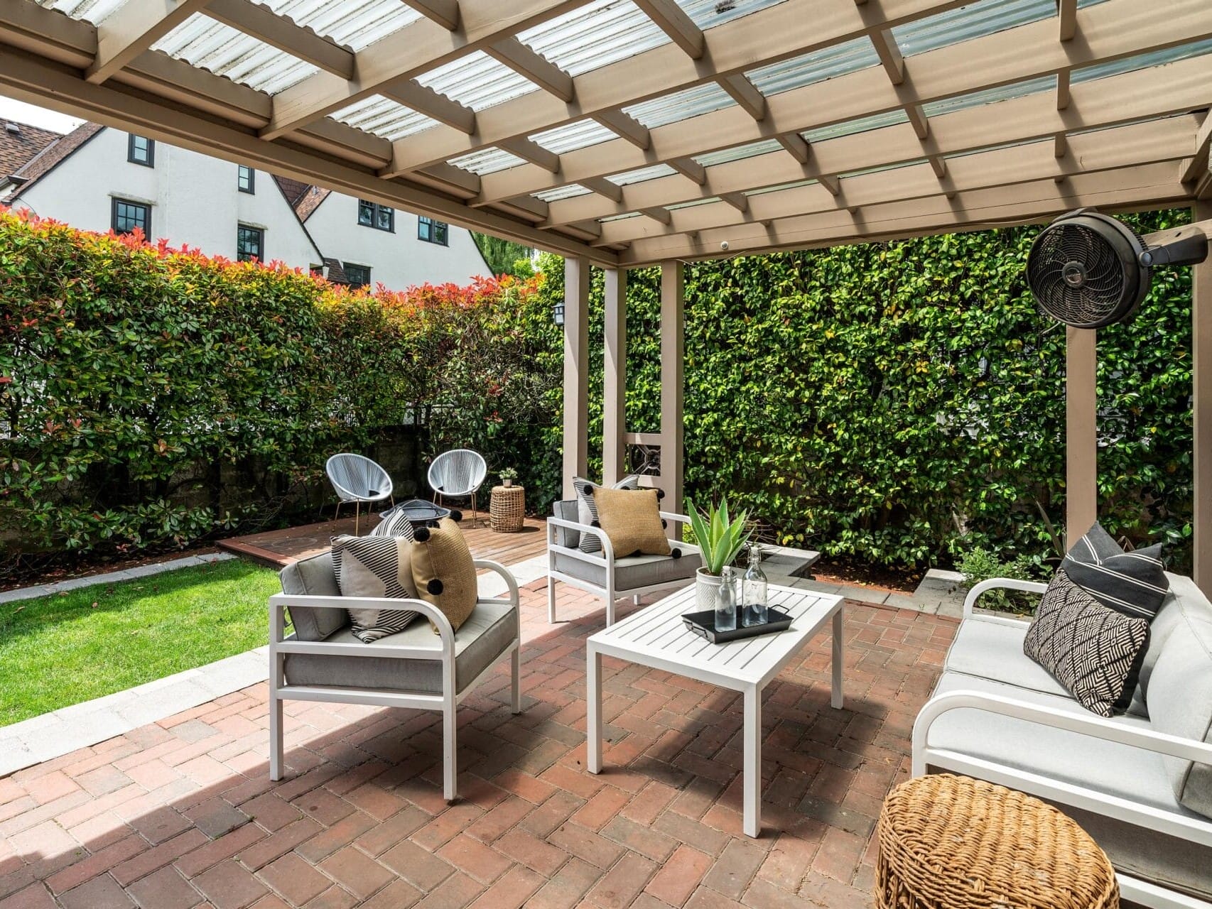 A cozy outdoor patio with white furniture under a wooden pergola. The seating area includes a coffee table and cushions, surrounded by lush green hedges. Two chairs are placed on a wooden deck, and a wicker basket sits on the brick floor.