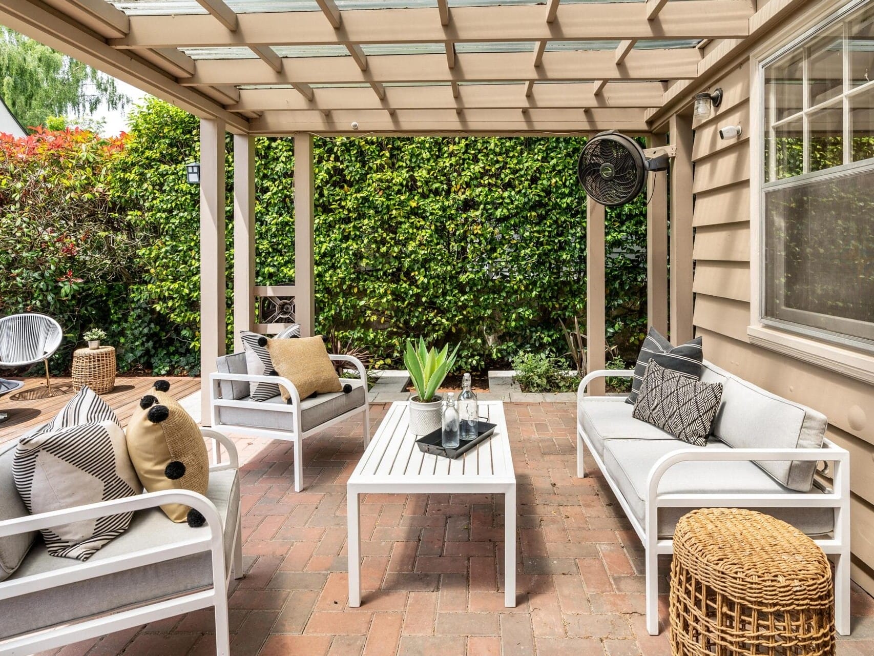 A cozy outdoor patio features white cushioned chairs and a matching table on a brick floor. The space is surrounded by green foliage. There are decorative pillows, a small plant on the table, and a wicker stool for added texture.