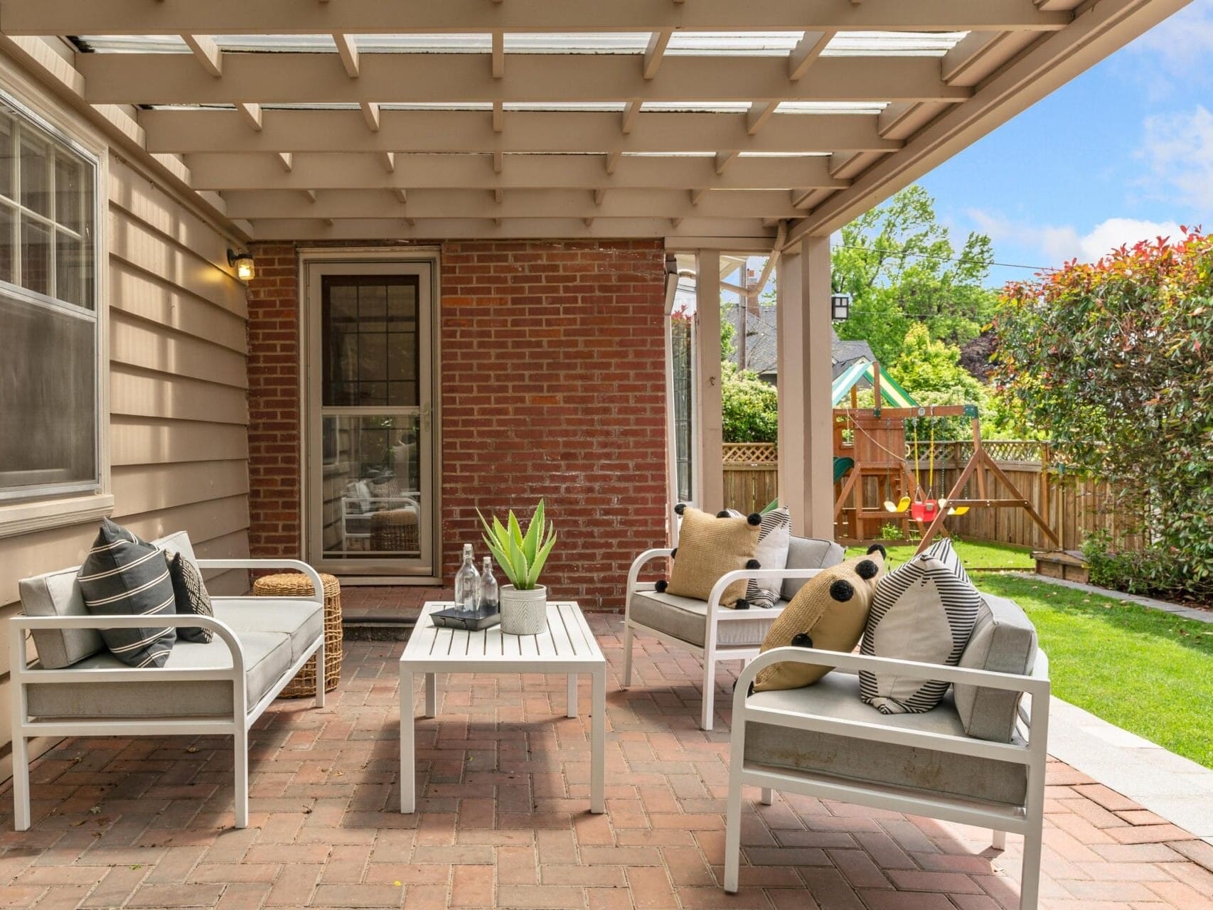 A cozy outdoor patio with a pergola, featuring white cushioned chairs and a coffee table on a brick floor. The space is decorated with potted plants and pillows. A lush garden with a playset is visible in the background.