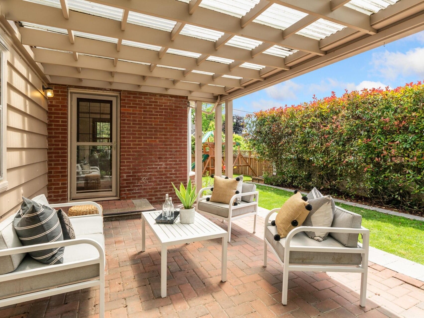 A cozy outdoor patio with a pergola covering, featuring a brick floor, white chairs with beige and black cushions, a small table with decorative items, and a lush hedge in the background. The scene is bright and inviting.