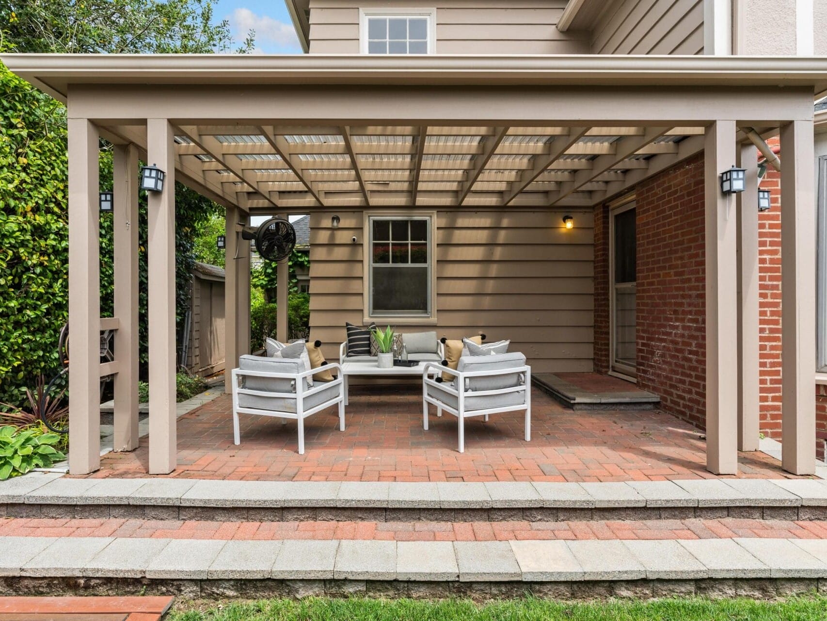 A cozy outdoor patio with a covered pergola features a seating area with four chairs and a coffee table on a brick floor. The house is painted beige with brick accents. Lush greenery surrounds the space, creating a relaxing atmosphere.