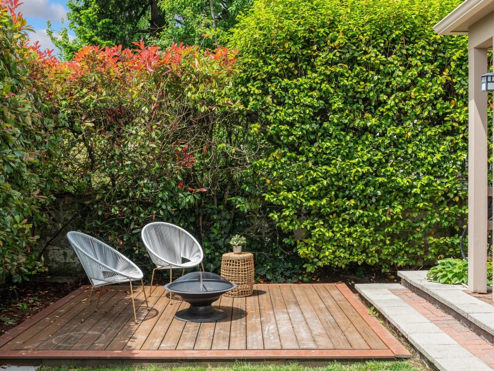 A wooden deck with two modern white chairs and a small table is set amidst lush green bushes. A round fire pit is placed in front of the chairs, providing a cozy outdoor seating area. The scene is tranquil and inviting.