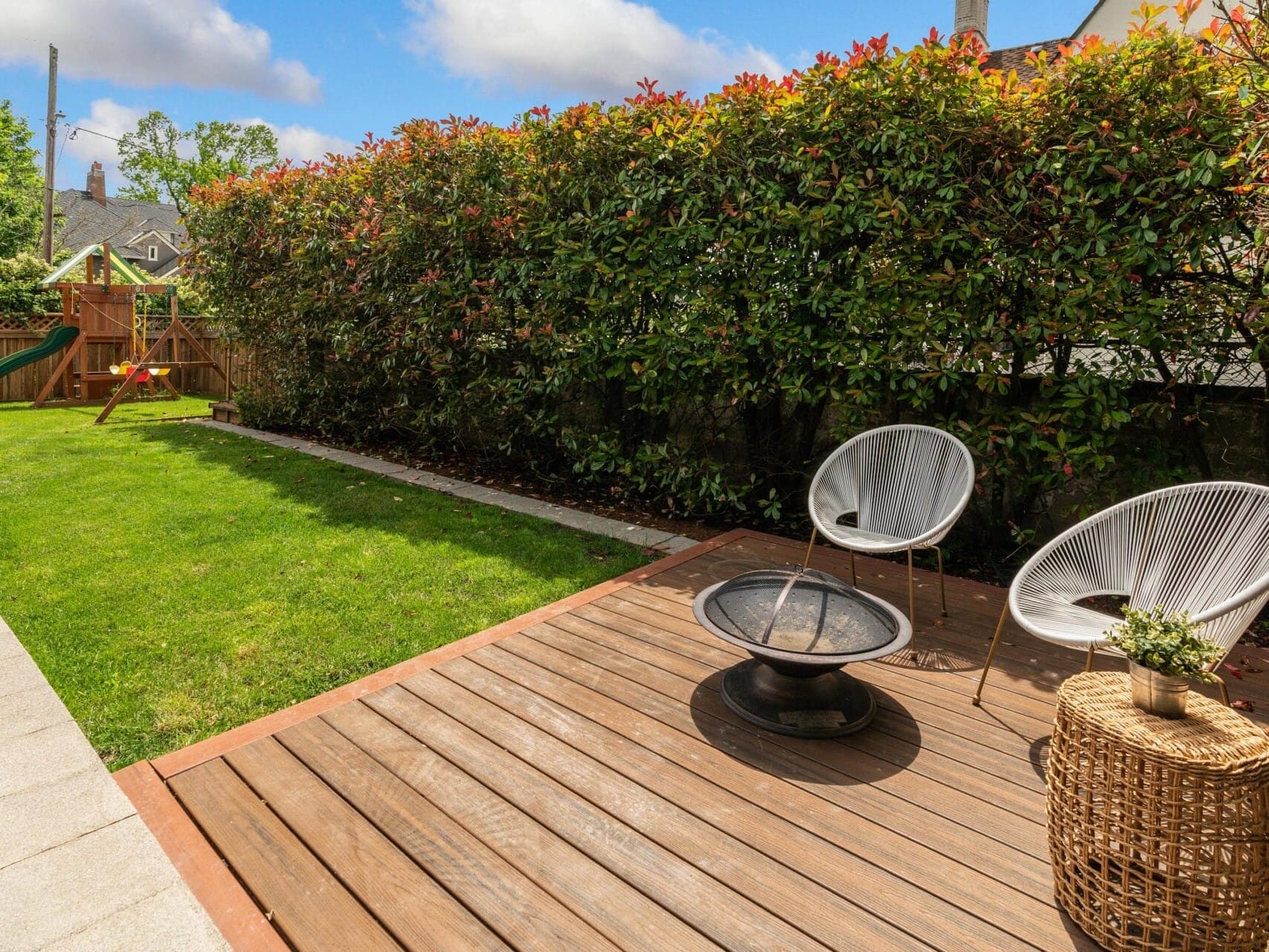 A backyard with a wooden deck featuring two modern chairs and a fire pit. Adjacent is a grassy lawn with a playset, surrounded by a tall hedge. A wicker table with a plant adds a decorative touch. Bright, clear skies overhead.