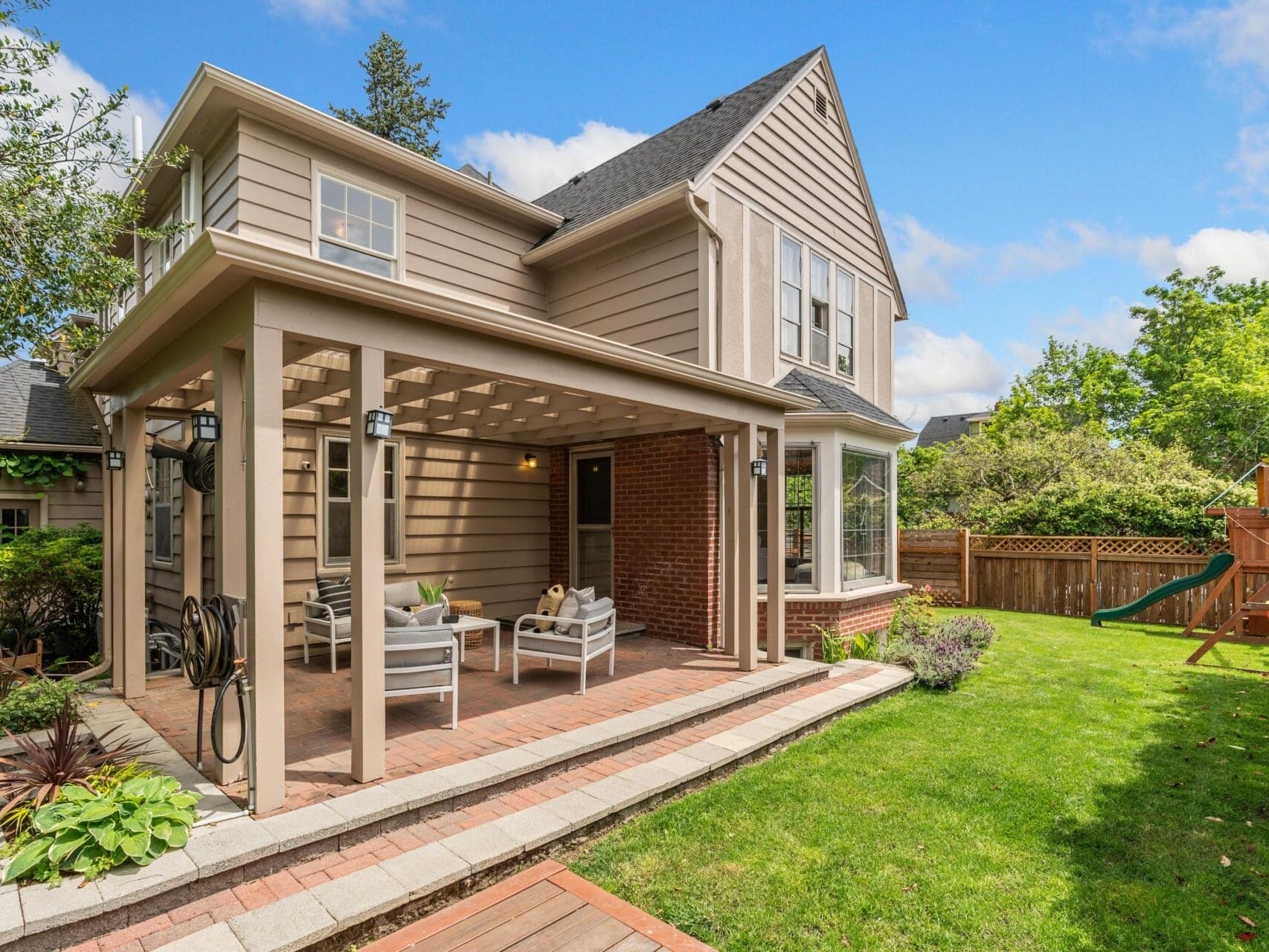 Two-story house with a covered patio featuring outdoor seating. The yard has a well-maintained lawn, a playset, and is bordered by a wooden fence. The sky is blue with clouds, and trees are visible in the background.