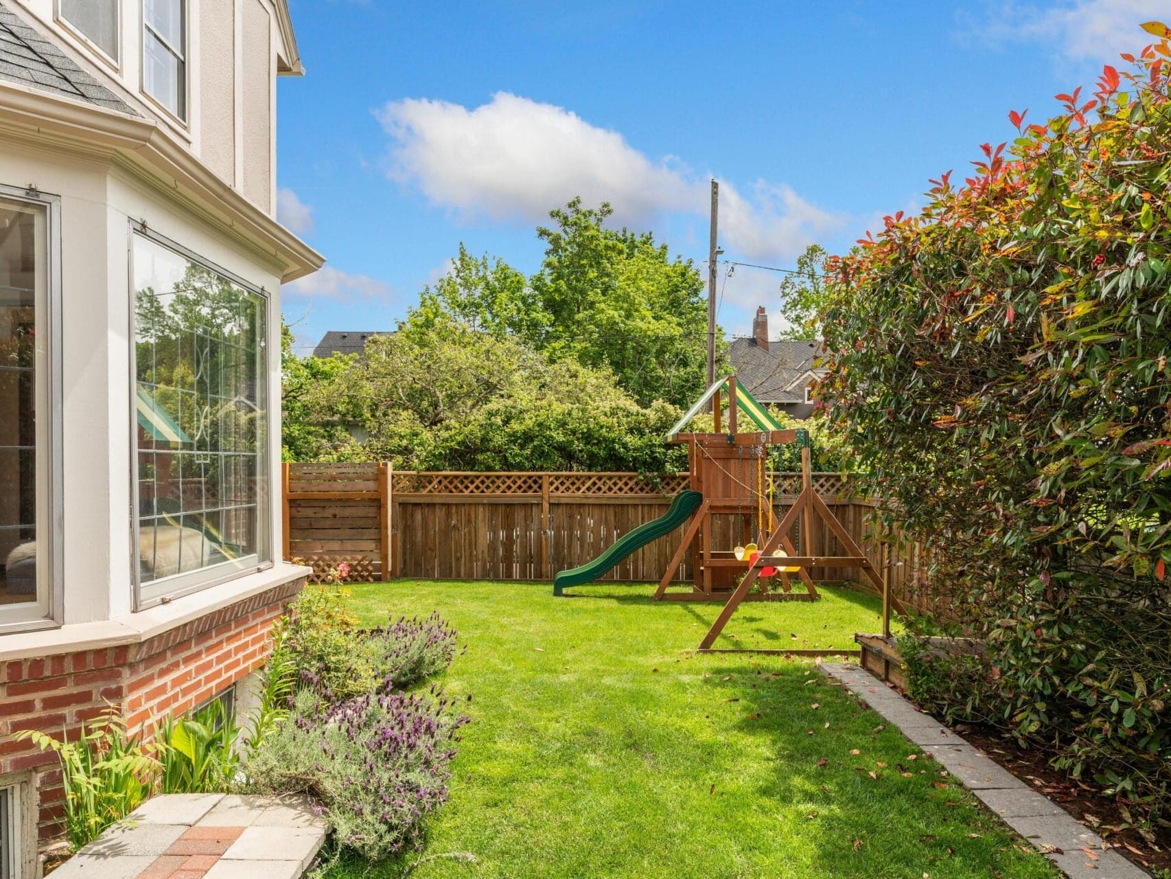 A backyard with a green slide and wooden swing set. The lawn is surrounded by a wooden fence with bushes and trees. Part of a brick house with large windows is visible on the left. The sky is clear with a few clouds.