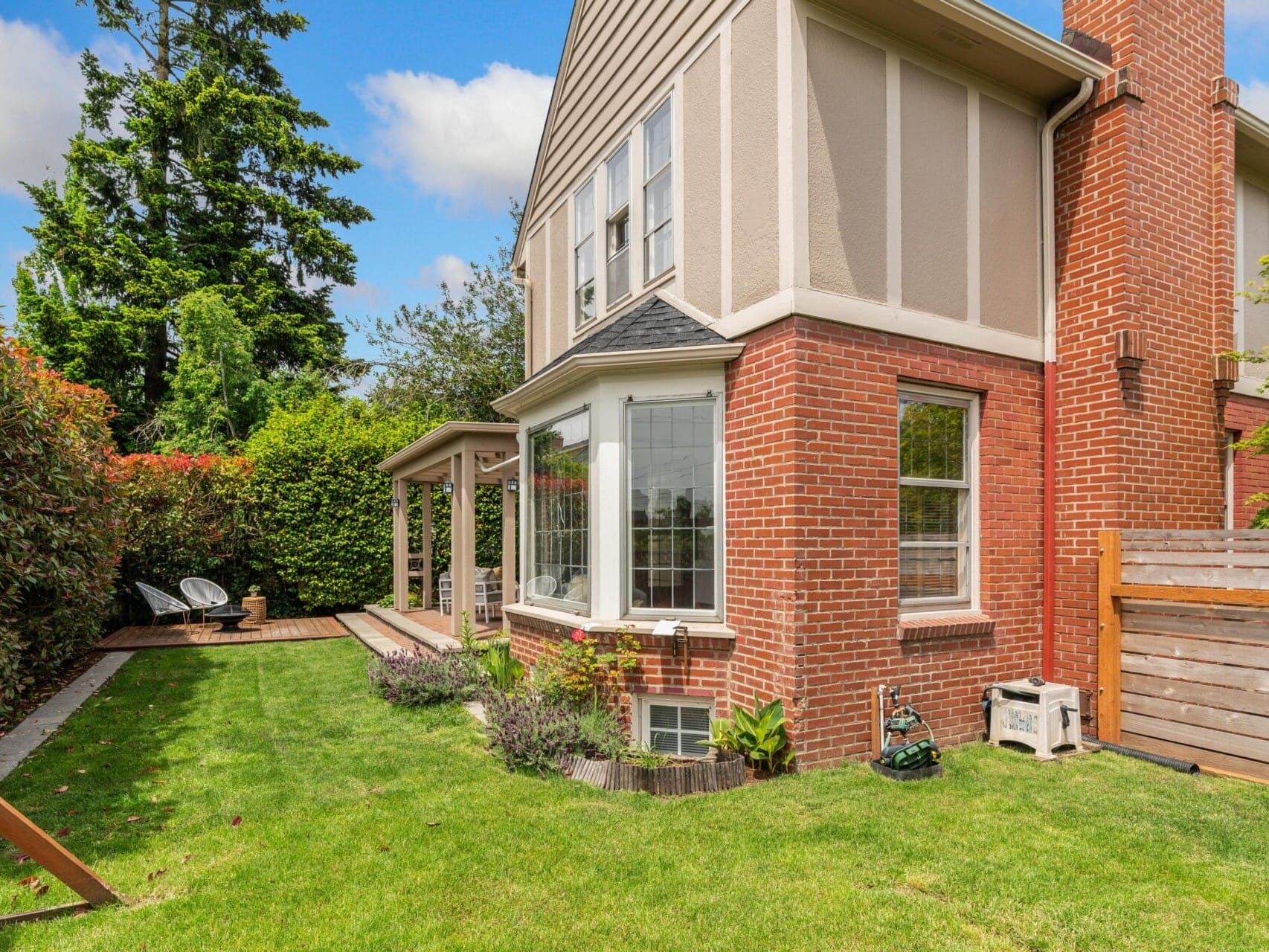 A two-story brick house with a well-manicured lawn and a small patio area on the side. The patio has two chairs and a wooden fence borders the yard, with tall trees and bushes providing a green backdrop. A sunny day with a blue sky and clouds.