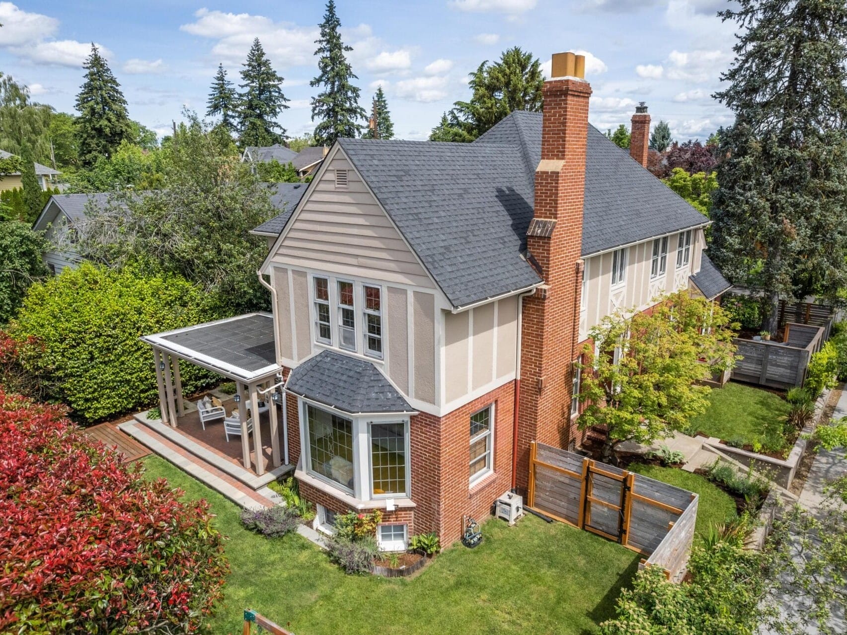 Aerial view of a charming two-story house with a mix of brick and beige siding. It features a covered porch, bay windows, and a well-maintained garden. Tall trees surround the property, and a wooden fence borders the backyard.