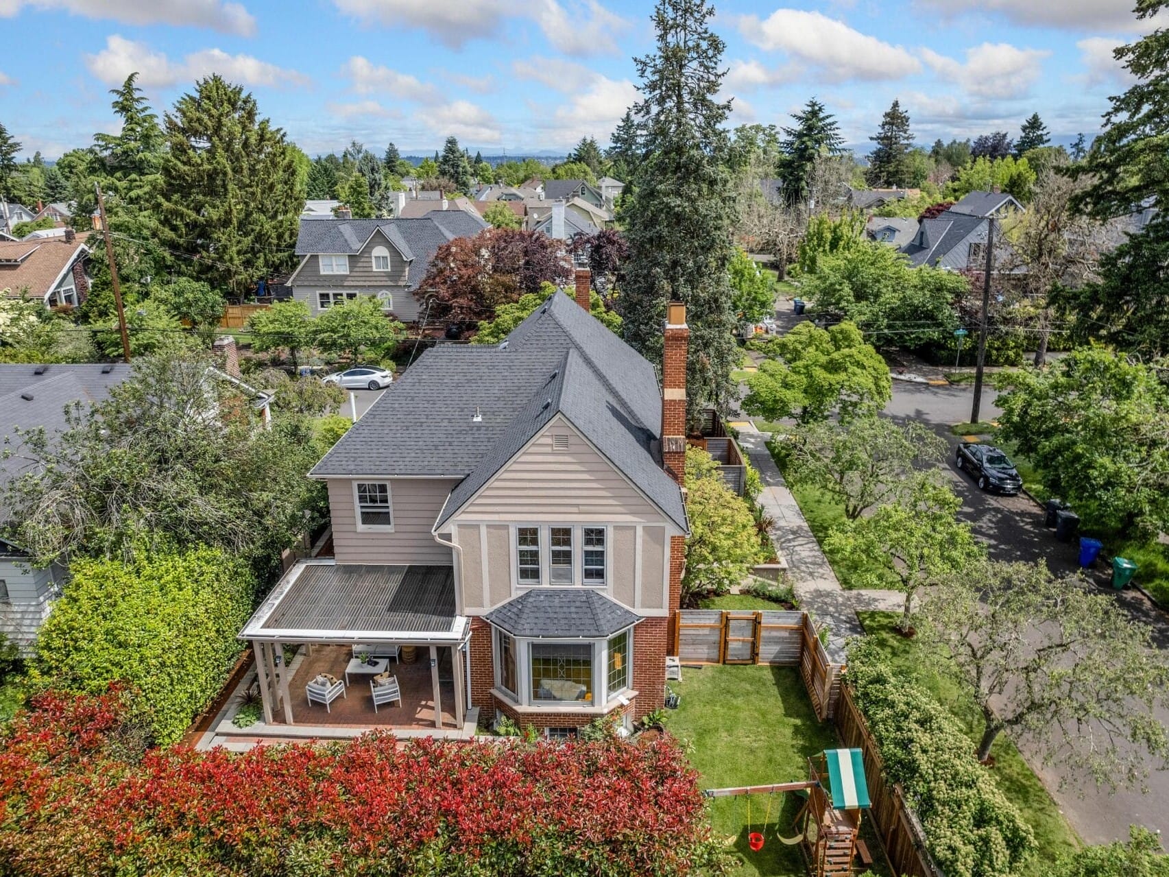 Aerial view of a suburban two-story house with a dark roof and a backyard. The yard features a small swing set and is bordered by greenery. The neighborhood has lush trees and similar houses visible in the distance under a partly cloudy sky.