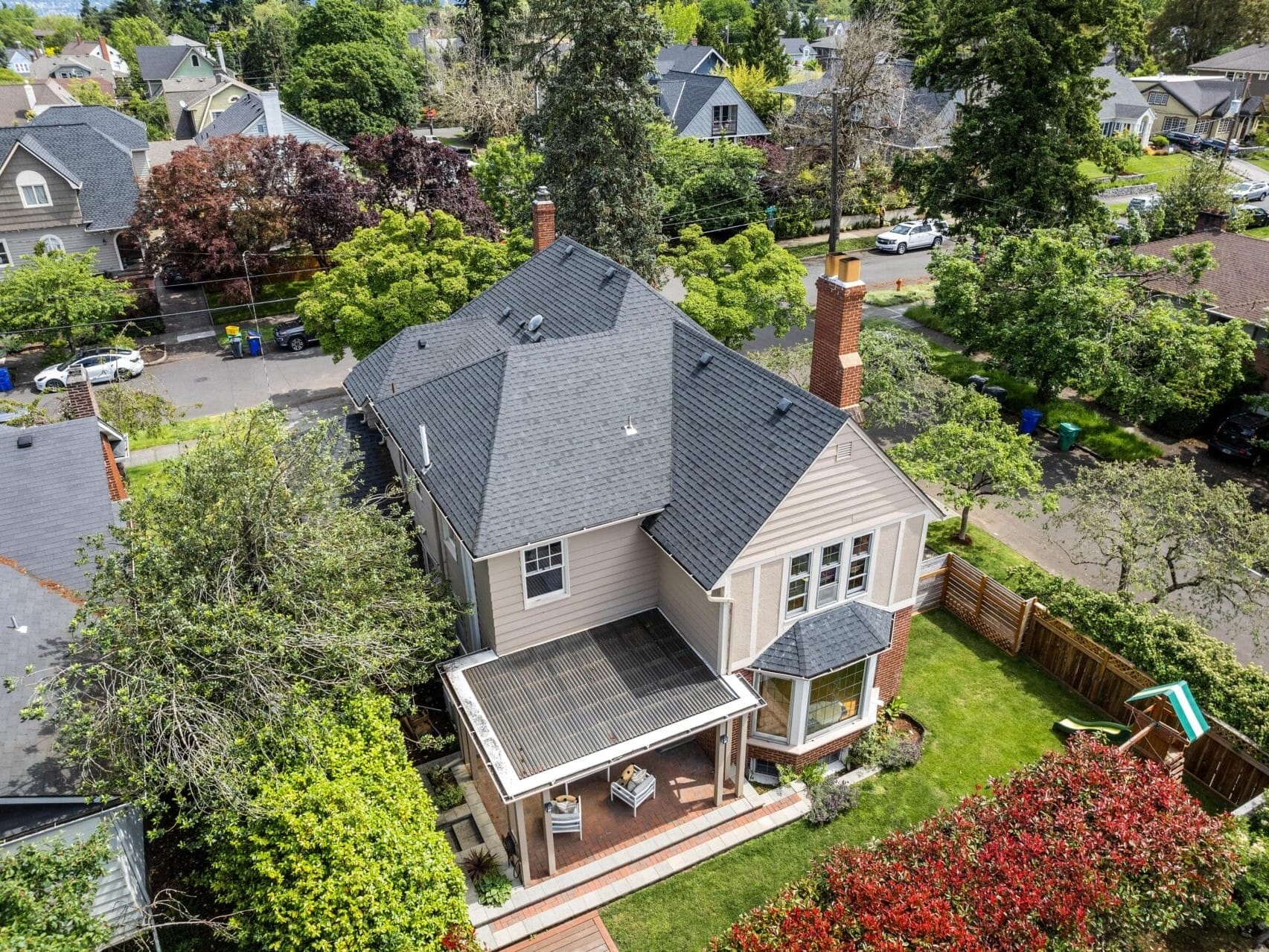 Aerial view of a suburban neighborhood featuring a large house with a dark roof, a covered patio, and a neatly manicured garden. Surrounding homes and tree-lined streets are visible, creating a tranquil and leafy residential scene.