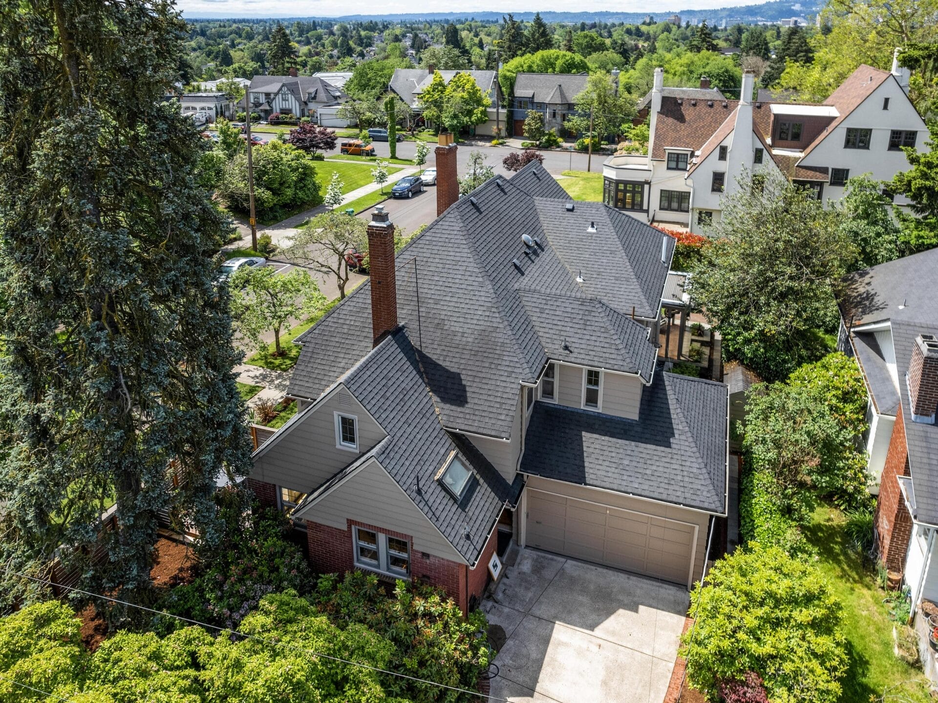 Aerial view of a suburban house with a gable roof, surrounded by trees and greenery. The driveway leads to a two-car garage. Nearby are similar homes and a tree-lined street, with a backdrop of a cityscape and distant hills.