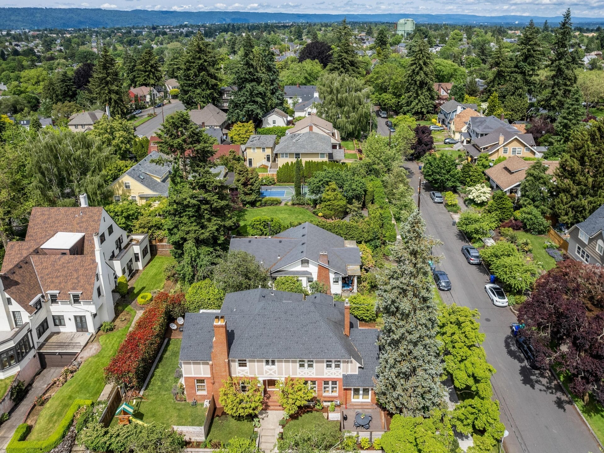 Aerial view of a suburban neighborhood with tree-lined streets and houses with backyards. The area is lush and green, with a variety of homes surrounded by trees. A clear sky and distant hills are visible on the horizon.