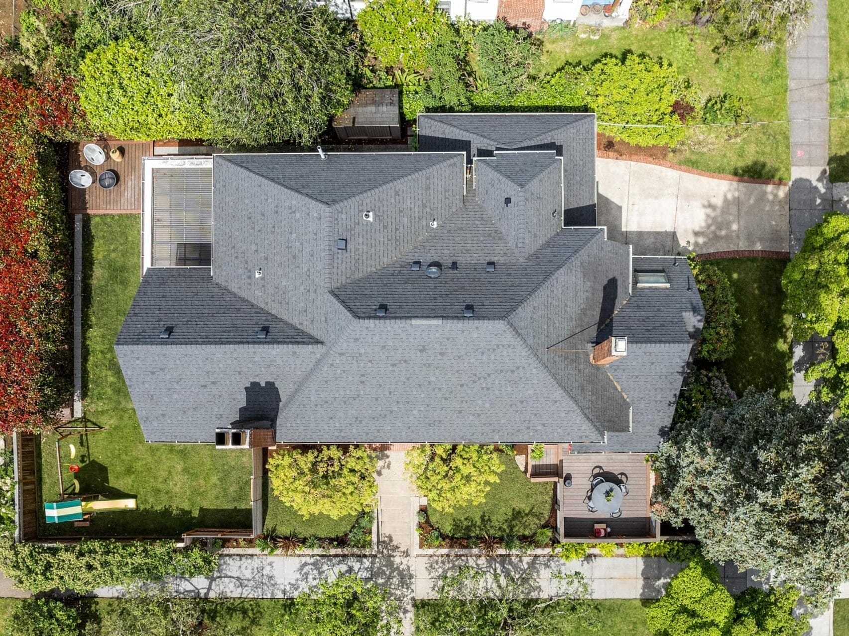 Aerial view of a large house with a dark roof, surrounded by a well-maintained garden. A paved path leads to the entrance. A small patio with furniture is visible on the right, and a childrens slide is on the left in a fenced yard.