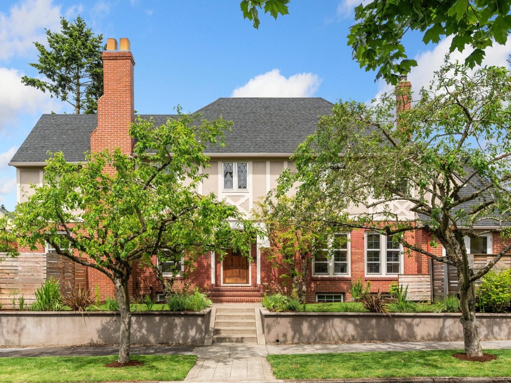 A two-story house with a red brick facade, a dark shingled roof, and a central wooden door. The front yard is landscaped with two leafy trees, bushes, and a manicured lawn. The sky is blue with scattered clouds.