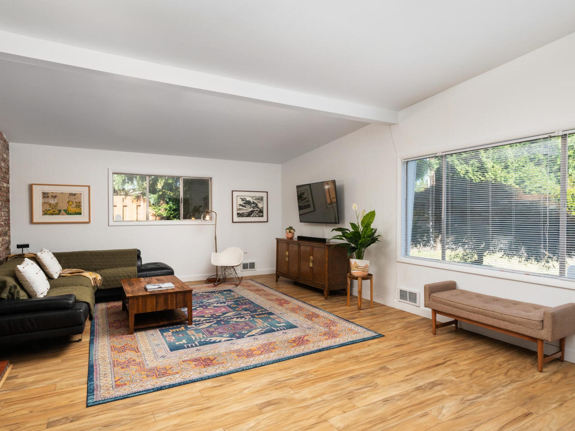 A spacious living room with wooden flooring, a large rug, two sofas, a coffee table, and a wall-mounted TV. Large window with blinds on the right and a green plant near a wooden cabinet. Decor includes framed artwork and cushions.