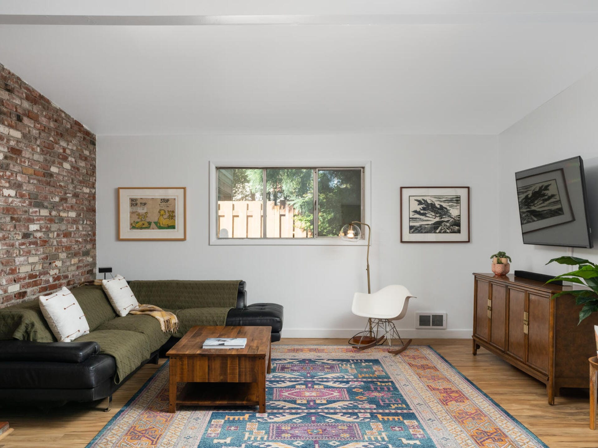 A cozy living room with a brick accent wall, a black sectional sofa, a wooden coffee table, and a colorful rug. A large window, wall art, a TV, and various plants decorate the space. A floor lamp and a chair add to the modern decor.