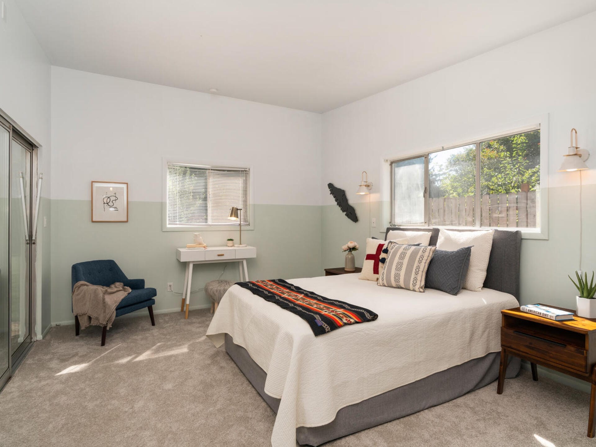 A cozy bedroom with a large bed featuring a white and patterned blanket. Theres a navy armchair and a white desk with a chair by a window. Two bedside tables hold lamps. Natural light fills the room through large windows.