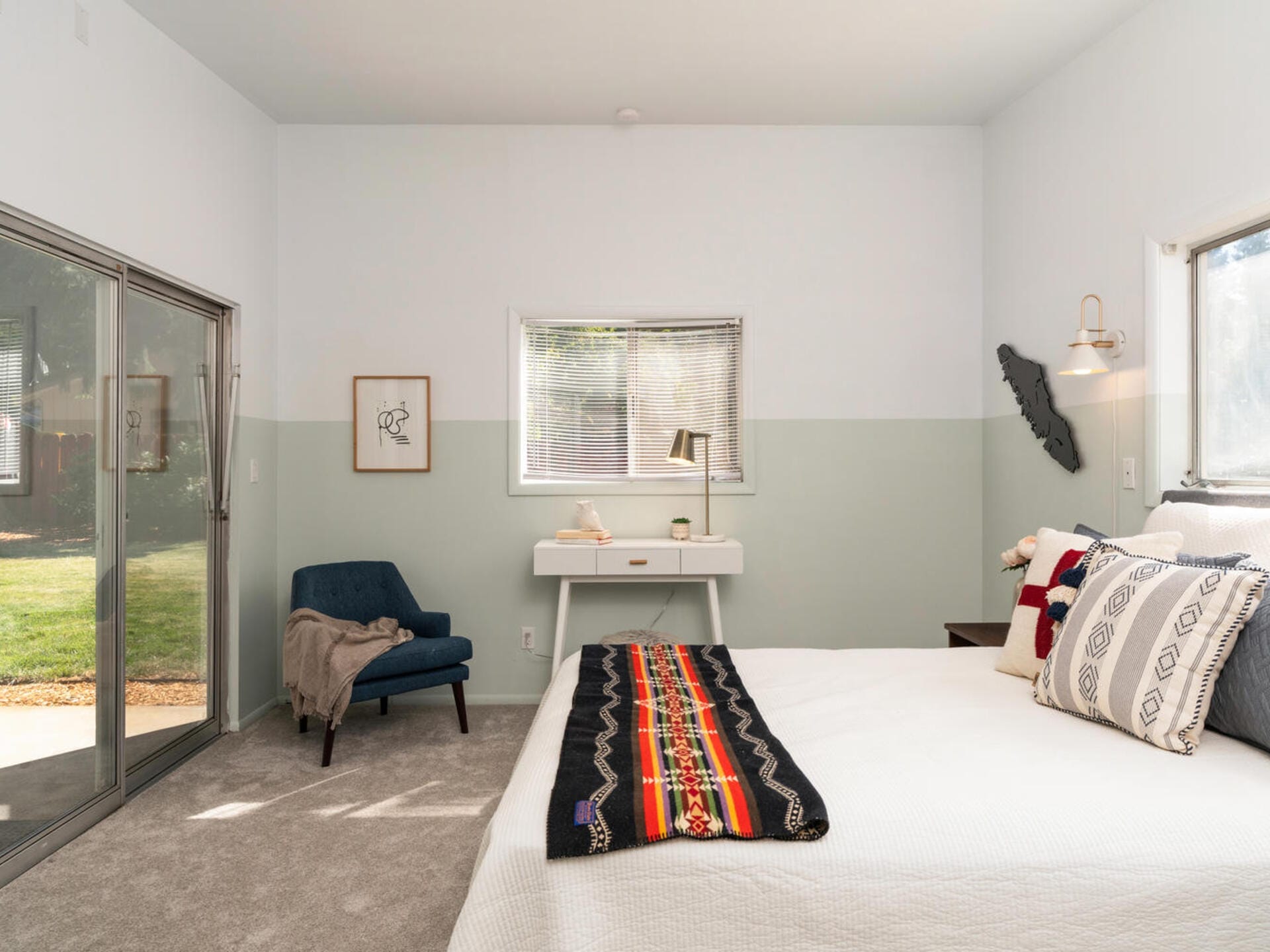 A cozy bedroom with a large bed featuring patterned throws and pillows. Theres a small desk and lamp under a window, an armchair with a blanket, and sliding glass doors leading to a garden. Soft, neutral tones create a serene atmosphere.