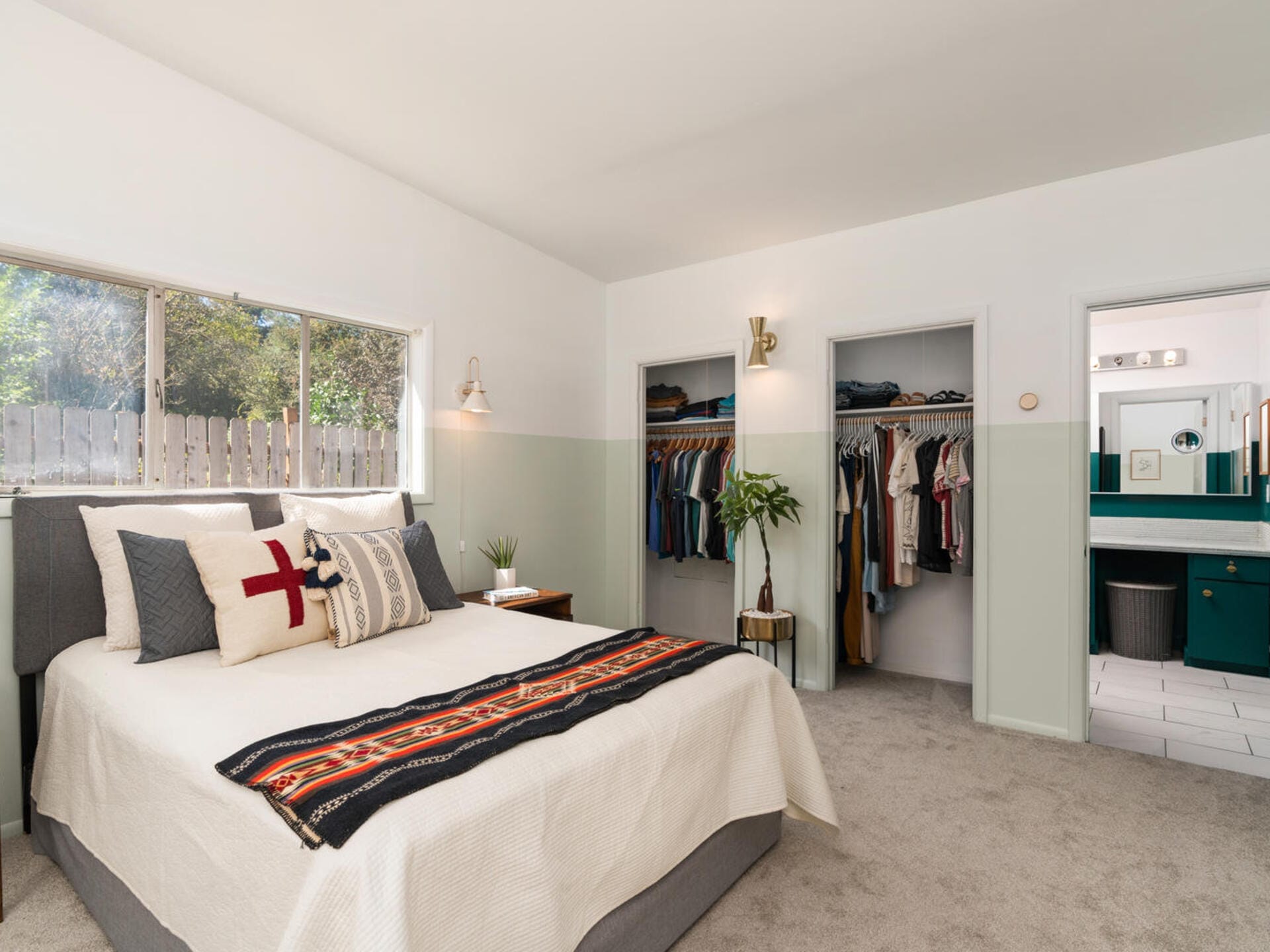 A cozy bedroom with a bed, decorative pillows, and a patterned throw. A partially open closet shows neatly arranged clothes. A plant sits by the closet, and a window brings in natural light. A bathroom is visible in the background.