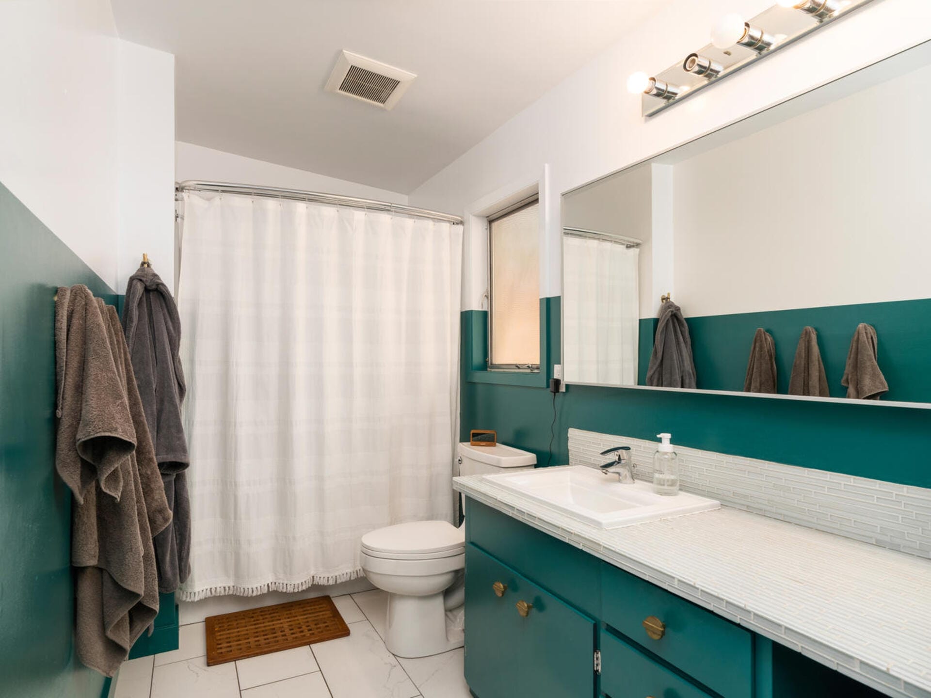 A bathroom with a white shower curtain, teal and white walls, and a large mirror above a sink. The vanity has teal cabinets and a white countertop. Towels hang on racks, and a toilet is positioned near the shower.