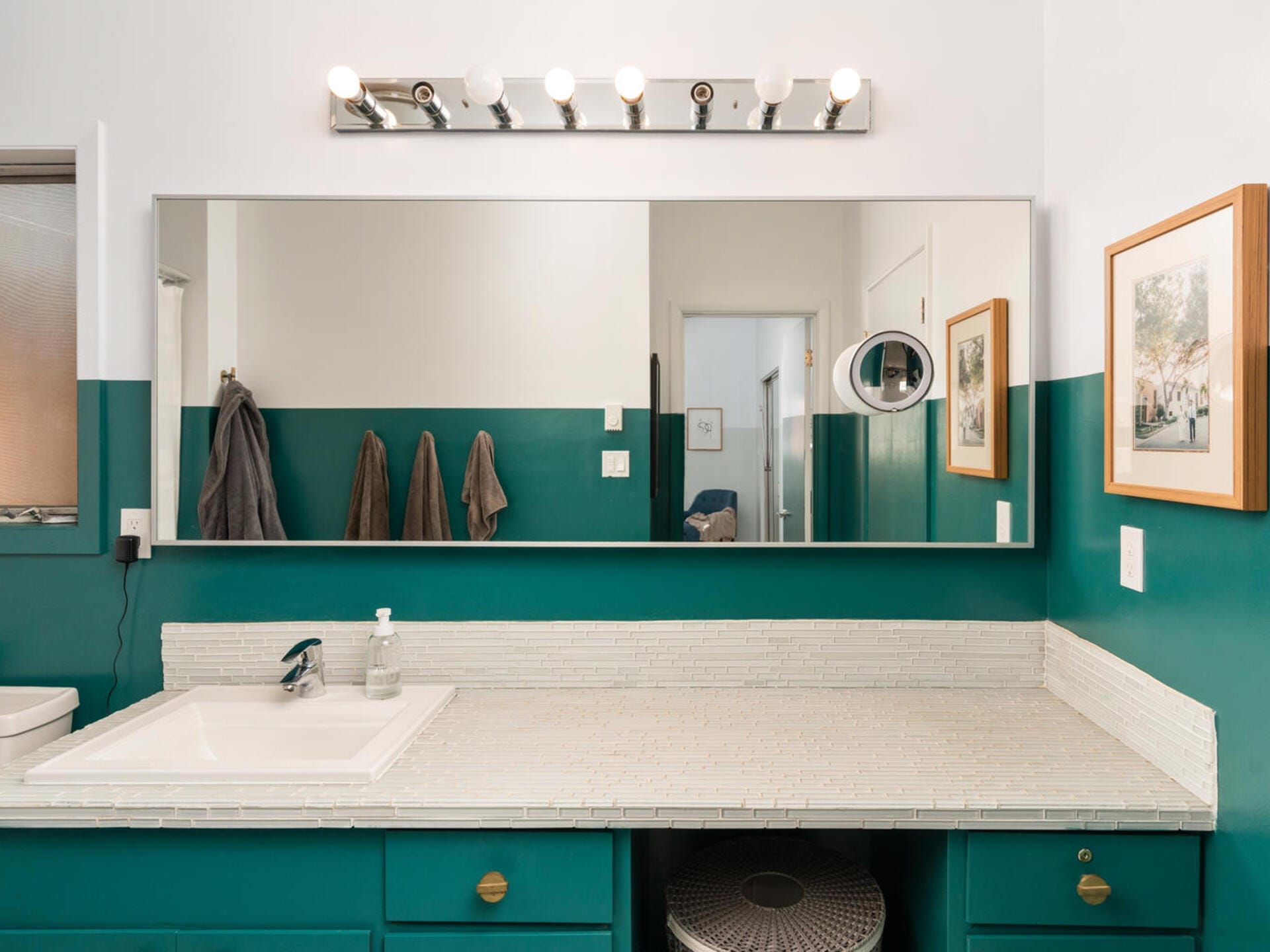 Bathroom with a long mirror above a white countertop, green cabinets, and a sink with a soap dispenser. Three towels hang on the wall. The walls are a mix of white and green, with framed artwork. Six-bulb vanity light fixture above the mirror.