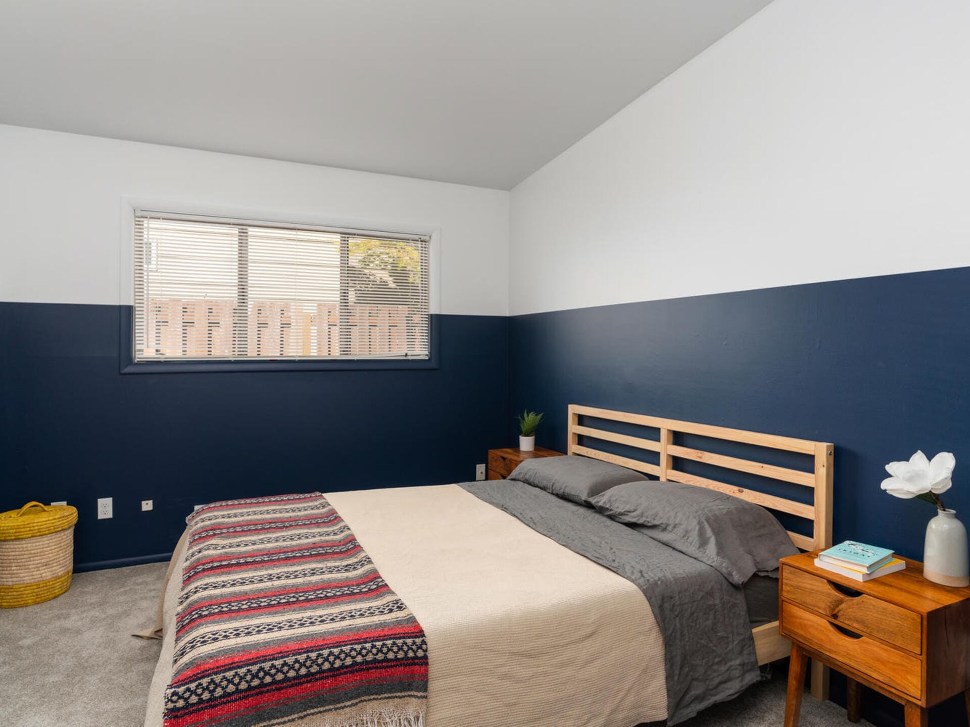 Minimalist bedroom with a wooden bed frame, gray bedding, and a patterned blanket. The walls are half white and half dark blue. Two wooden bedside tables hold a book, flower vase, and a small plant. A window with blinds lets in natural light.