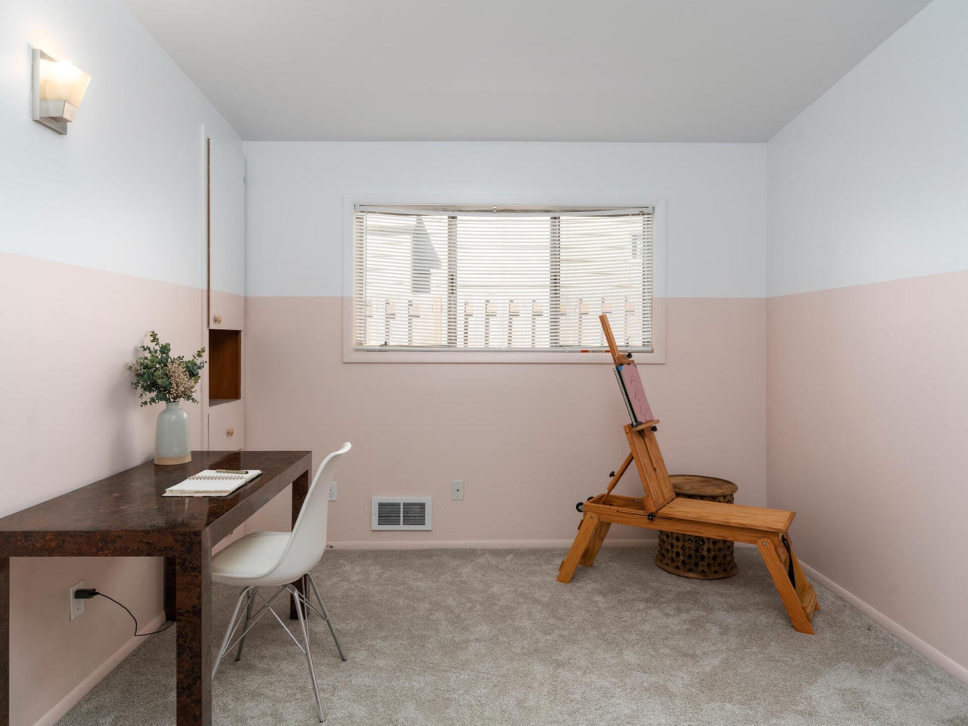 A minimalist room with a brown desk and white chair facing a window. An easel is positioned near the window, and a small plant decorates the desk. The walls are painted with a light beige lower half and a white upper half.