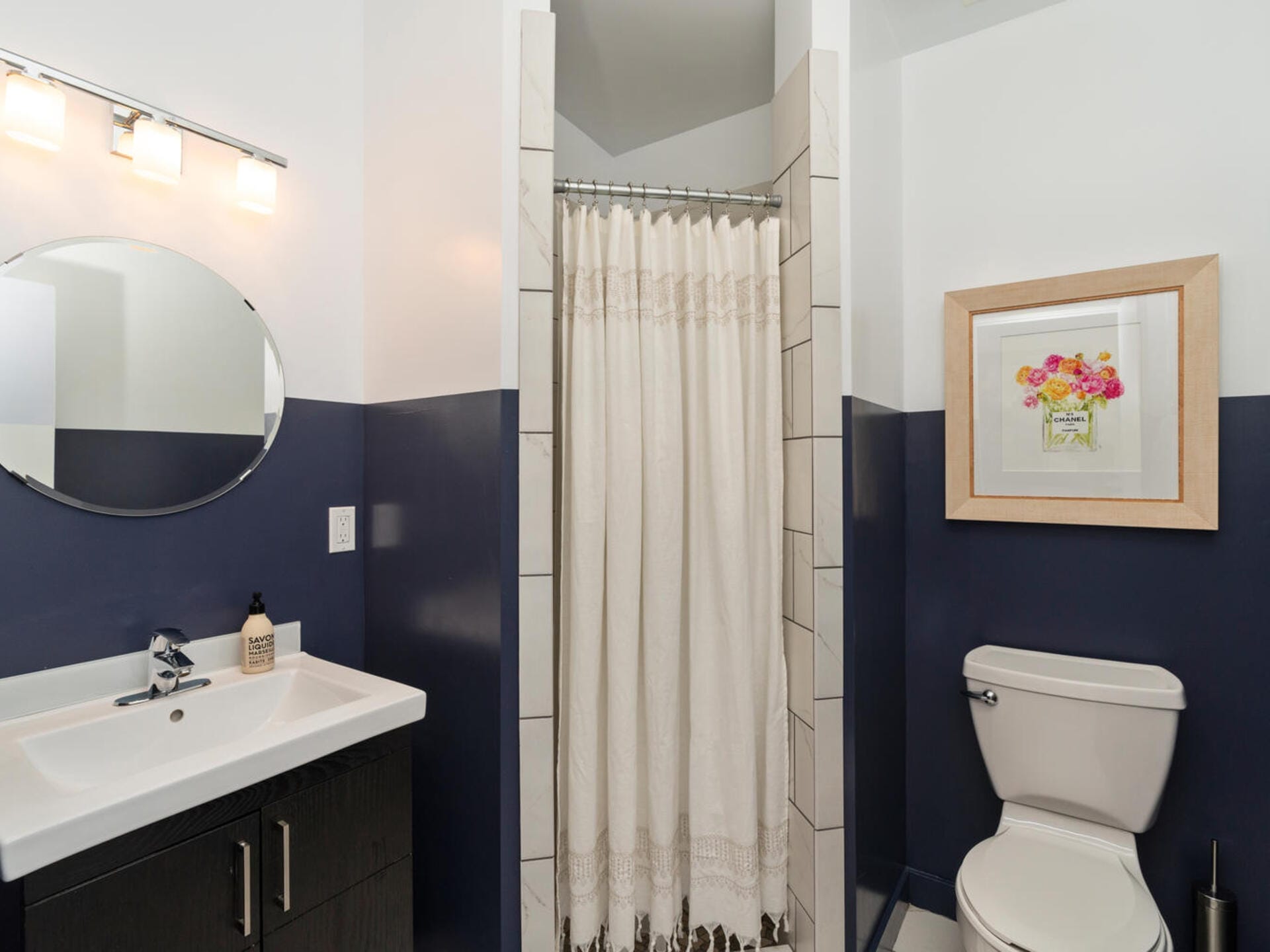 A modern bathroom with a white sink, a round mirror, a dark vanity, and a light above. A shower with a white curtain is in the corner, next to a toilet with a framed floral picture above it. The walls are half navy, half white.