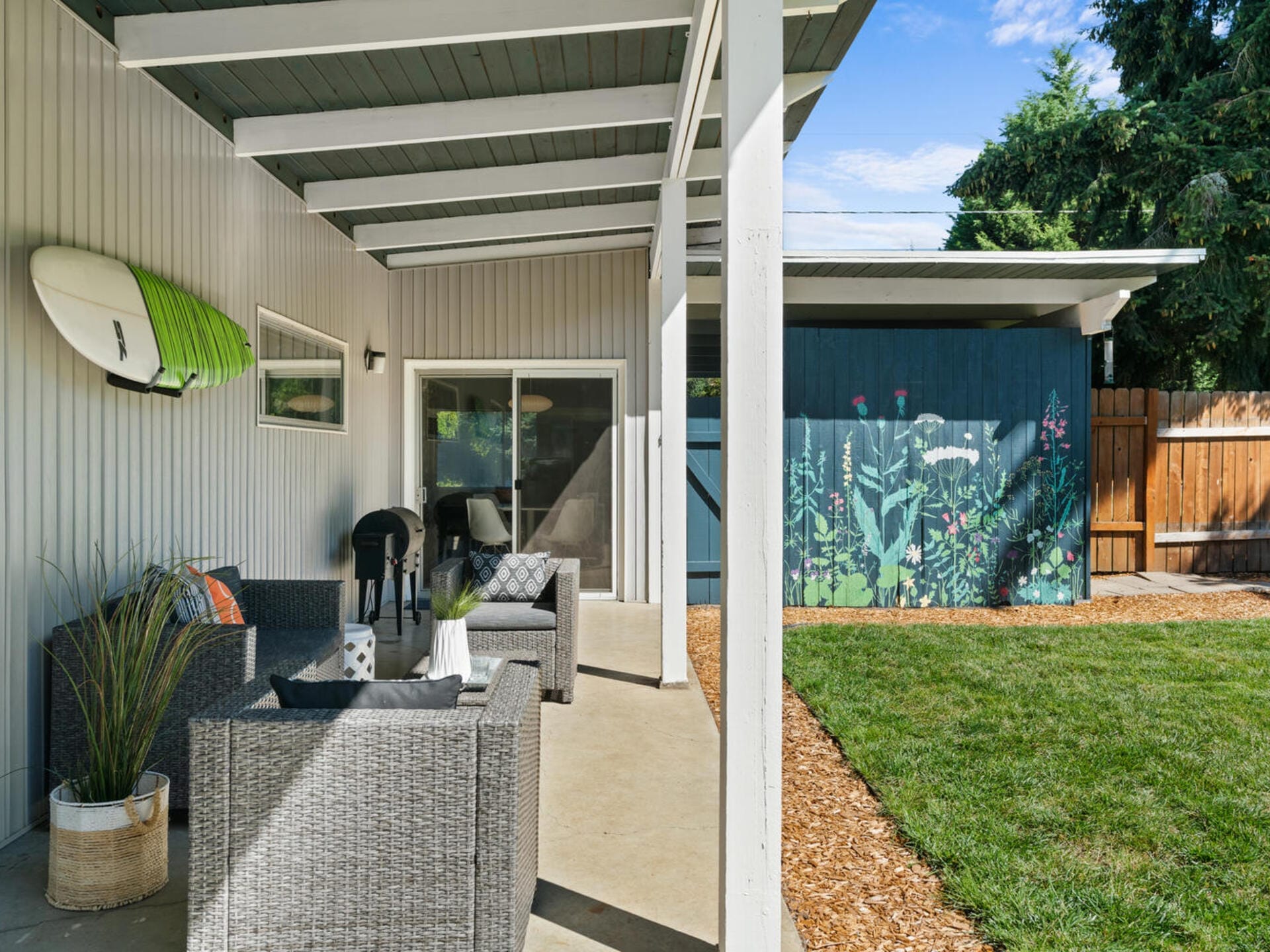 Covered patio area with gray wicker furniture, a small table, and cushions. A surfboard is mounted on the wall. The patio overlooks a backyard with a painted fence depicting plants and a grassy lawn. The sky is clear and sunny.