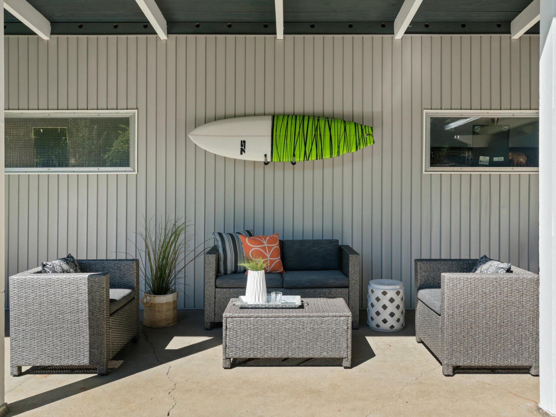 Cozy patio with gray wicker furniture, including a loveseat, two chairs, and a coffee table. A surfboard with green details hangs above, and two small windows are on either side. A plant and cushions add a touch of color.
