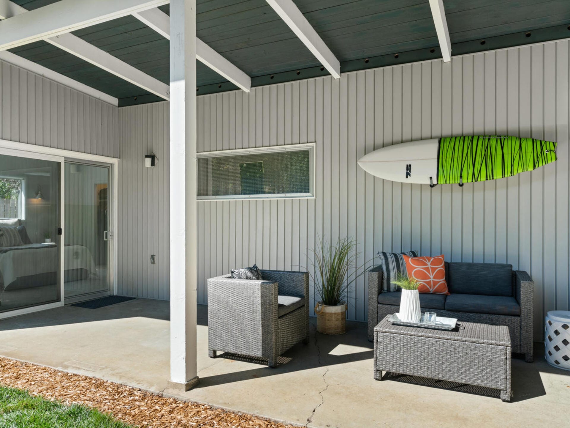 A covered patio featuring two gray wicker chairs with cushions, a matching coffee table, and a decorative surfboard mounted on the wooden wall. A small plant sits next to the chairs. The area leads into the house through sliding glass doors.