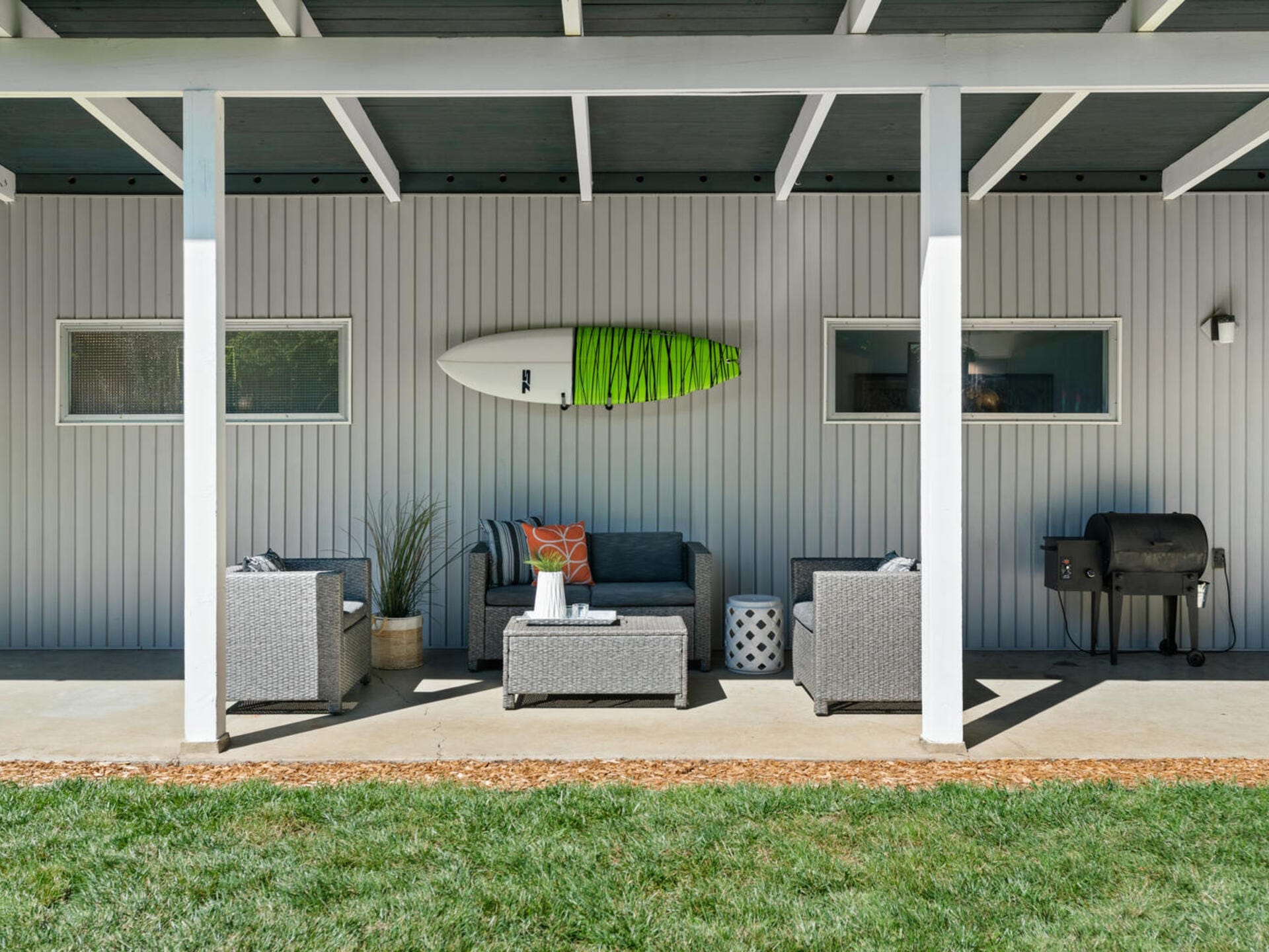 Outdoor patio with gray wicker furniture under a white pergola. A surfboard decorates the wall above the seating area. Two windows are visible, and a barbecue grill is on the right. Green grass edges the concrete patio floor.