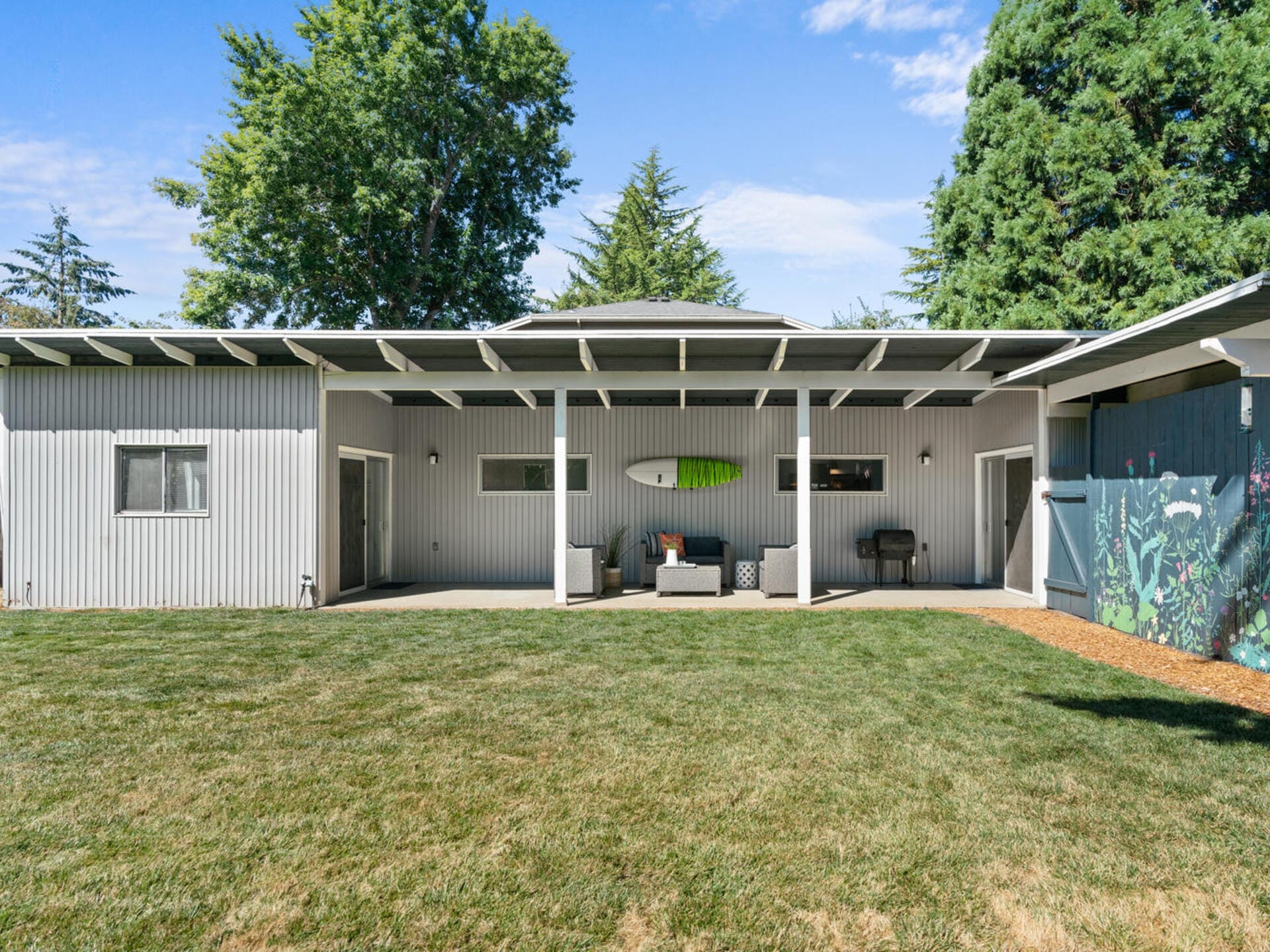 A modern, single-story building with a flat roof, light gray exterior, and large patio area. It has outdoor seating and a surfboard decor mounted on the wall. The yard is grassy and bordered by tall green trees.