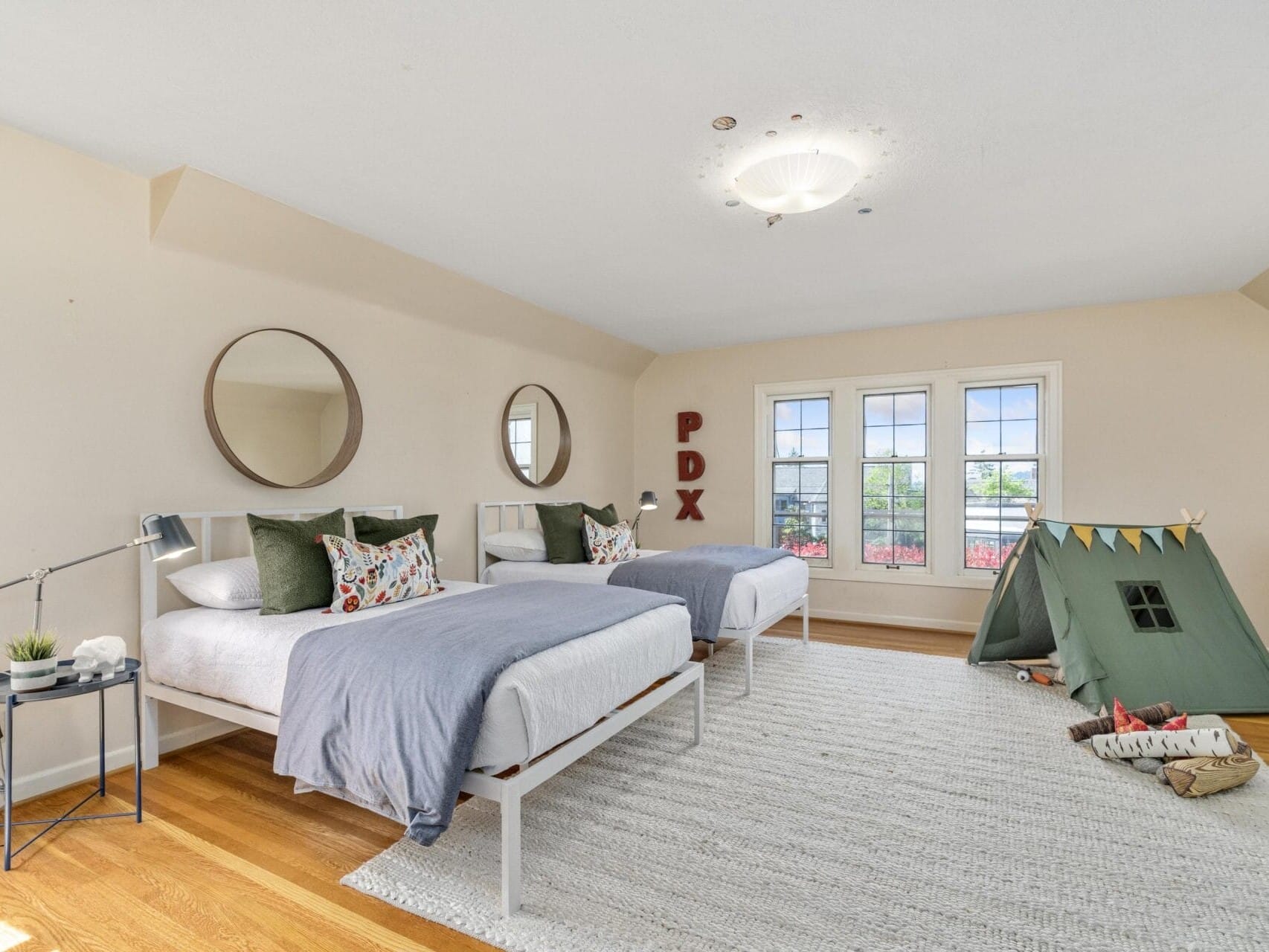 A spacious bedroom with two beds, round mirrors above each bed, a small tent with bunting, and a plush rug. Large windows let in natural light. Walls are beige, and the letters PDX are displayed in red on the wall.