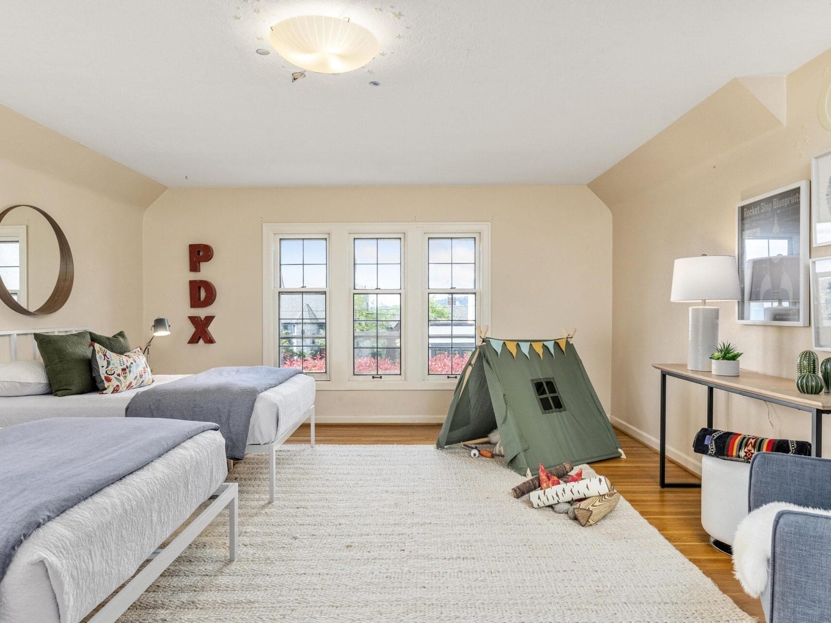 A cozy bedroom with two beds, a round mirror, and PDX letters on the wall. A green play tent is in the corner with cushions. Large windows let in natural light. A desk with a lamp and framed pictures is on the right. A light-colored rug covers the floor.