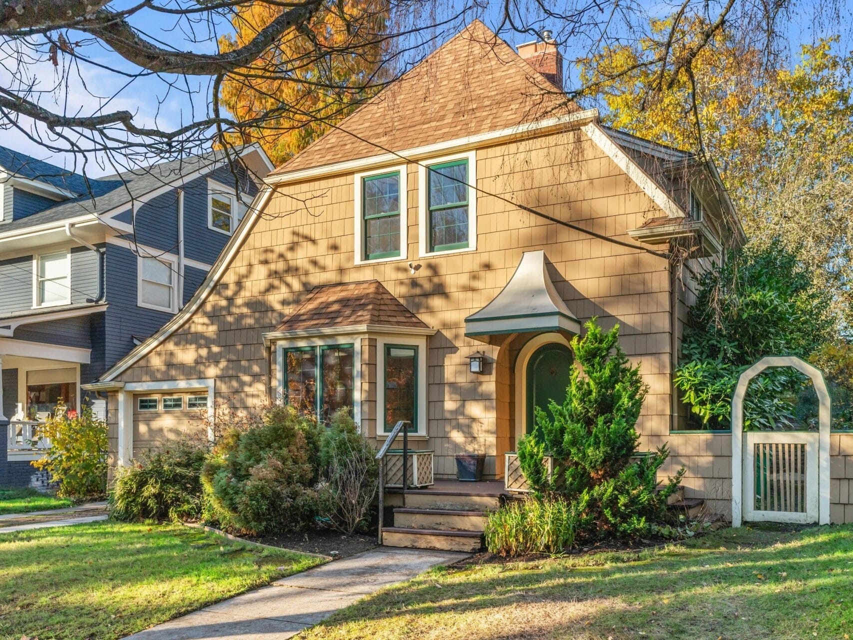 A charming brown house with a triangular roof and a curved green awning over the front door. Its set amidst autumn trees, and features a manicured lawn, garage, and a white fence with an arched gate. Neighboring house partially visible.