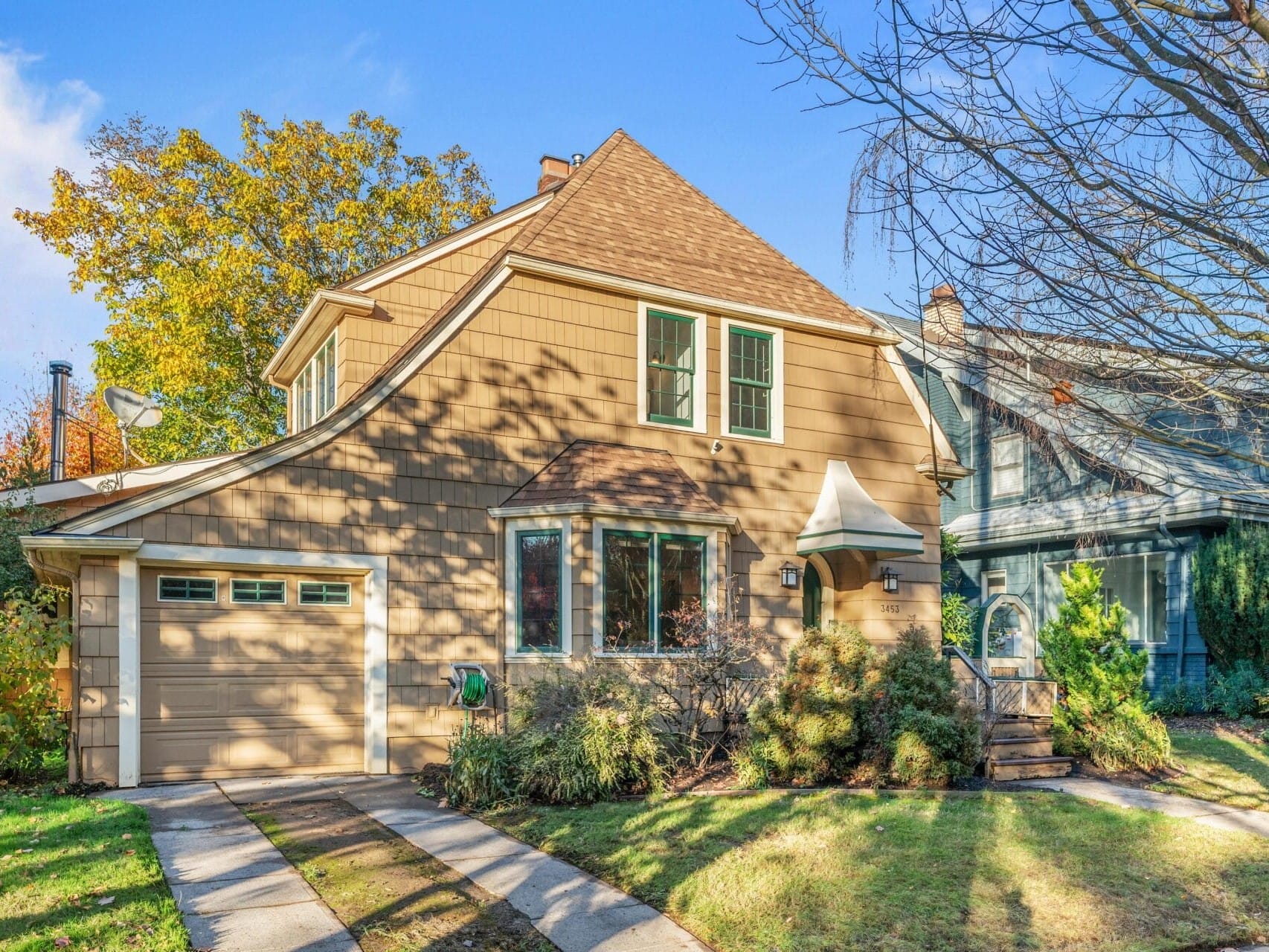 A charming two-story house with tan siding, surrounded by autumn trees. It features a one-car garage, a small front porch, and a well-maintained lawn. The sky is clear and blue, casting soft shadows on the home and yard.